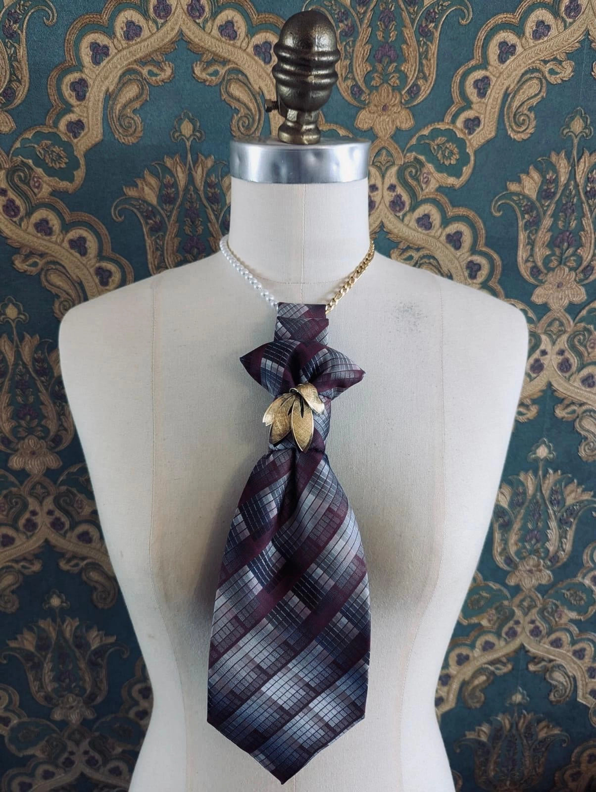 Broochella Montecristo cravatta vintage buurgundy and grey check handmade up cycled tie brooch with vintage brooch embellishment. It is pinned to a mannequin The background is green and gold damask wallpaper. 