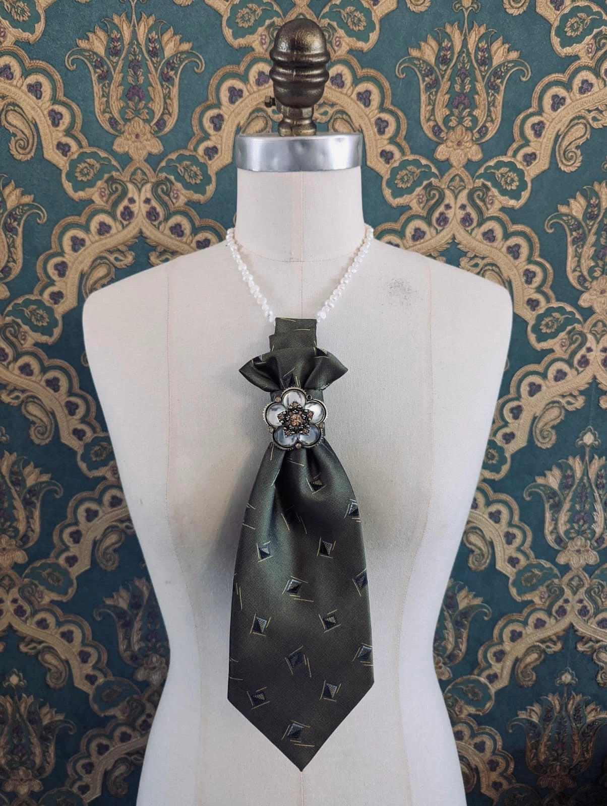 A Broochella Montecristo cravatta number 21 upcycled vintage tie brooch in taupe and black print, with a vintage flower brooch embellishment. It is pinned on a cream-coloured dress form mannequin. The background is green and gold damask wallpaper. 