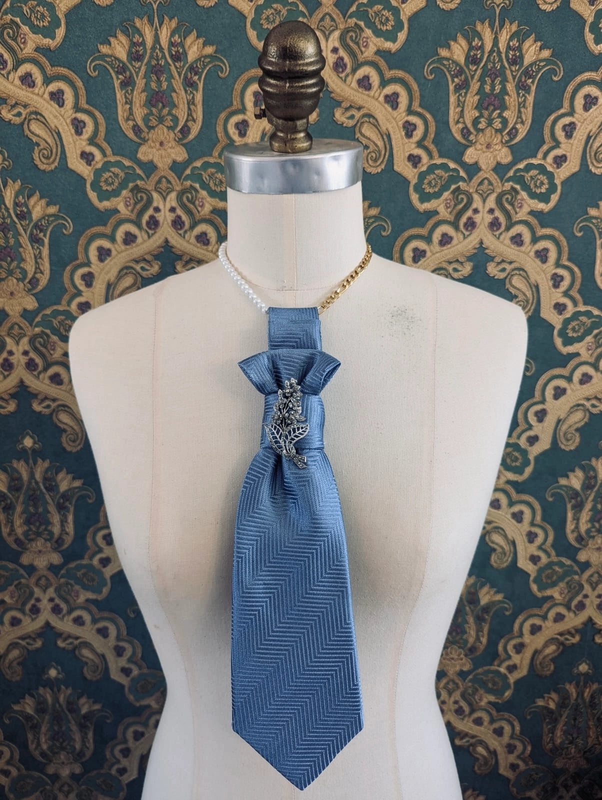 A Broochella Montecristo cravatta 13 blue herringbone patterned necktie styled on a cream-colored mannequin torso, featuring a decorative silver-toned brooch at the knot. The mannequin is set against a dark teal and gold damask patterned wallpaper background.