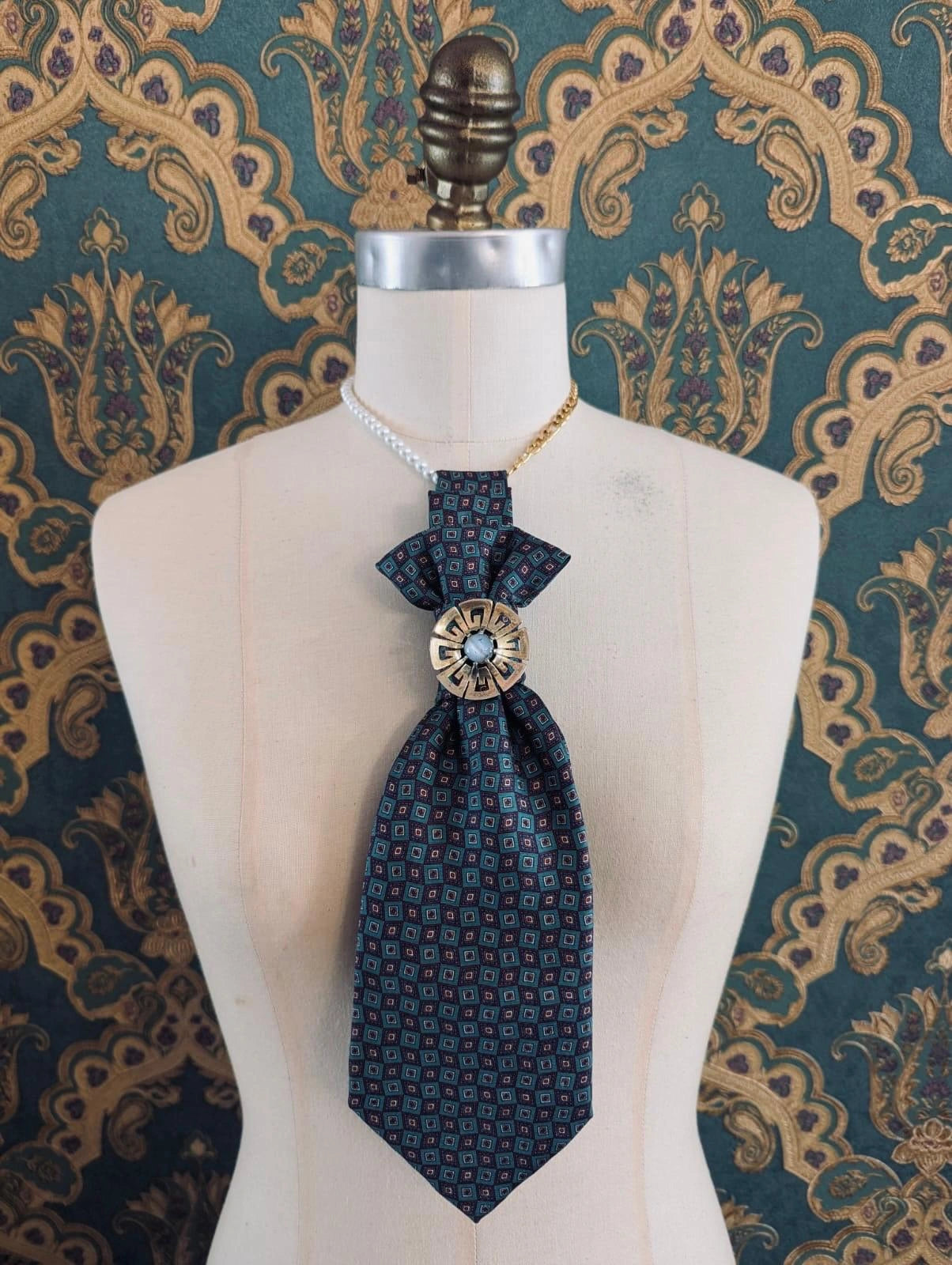 Broochella Montecristo cravatta vintage green check handmade up cycled tie brooch with vintage brooch embellishment. It is pinned to a mannequin The background is green and gold damask wallpaper. 