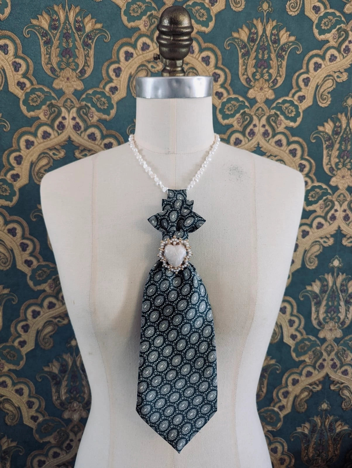 A Broochella Montecristo cravatta number 20 up-cycled vintage tie brooch in green and cream pattern, with a heart shaped brooch embellishment. It is pinned on a cream-coloured dress form mannequin. The background is green and gold damask wallpaper. 
