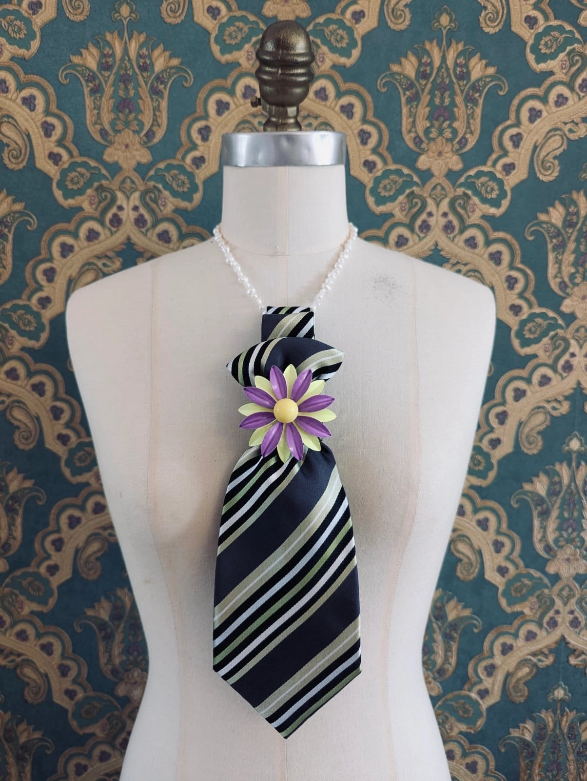 A Broochella Montecristo cravatta number 24  upcycled vintage tie brooch in black and white green, with a vintage brooch embellishment. It is pinned on a cream-coloured dress form mannequin. The background is green and gold damask wallpaper. 