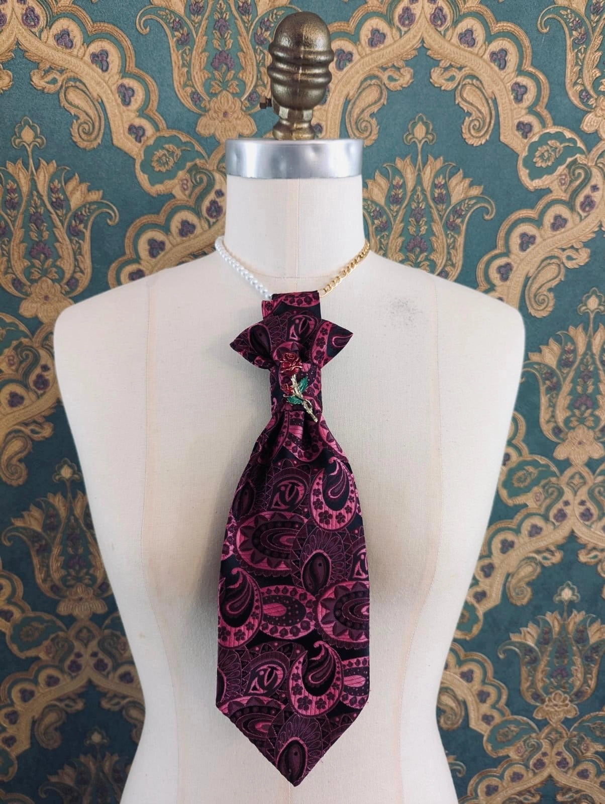 Broochella Montecristo cravatta vintage pink and black check handmade up cycled  pailsey tie brooch with vintage brooch embellishment. It is pinned to a mannequin The background is green and gold damask wallpaper. 