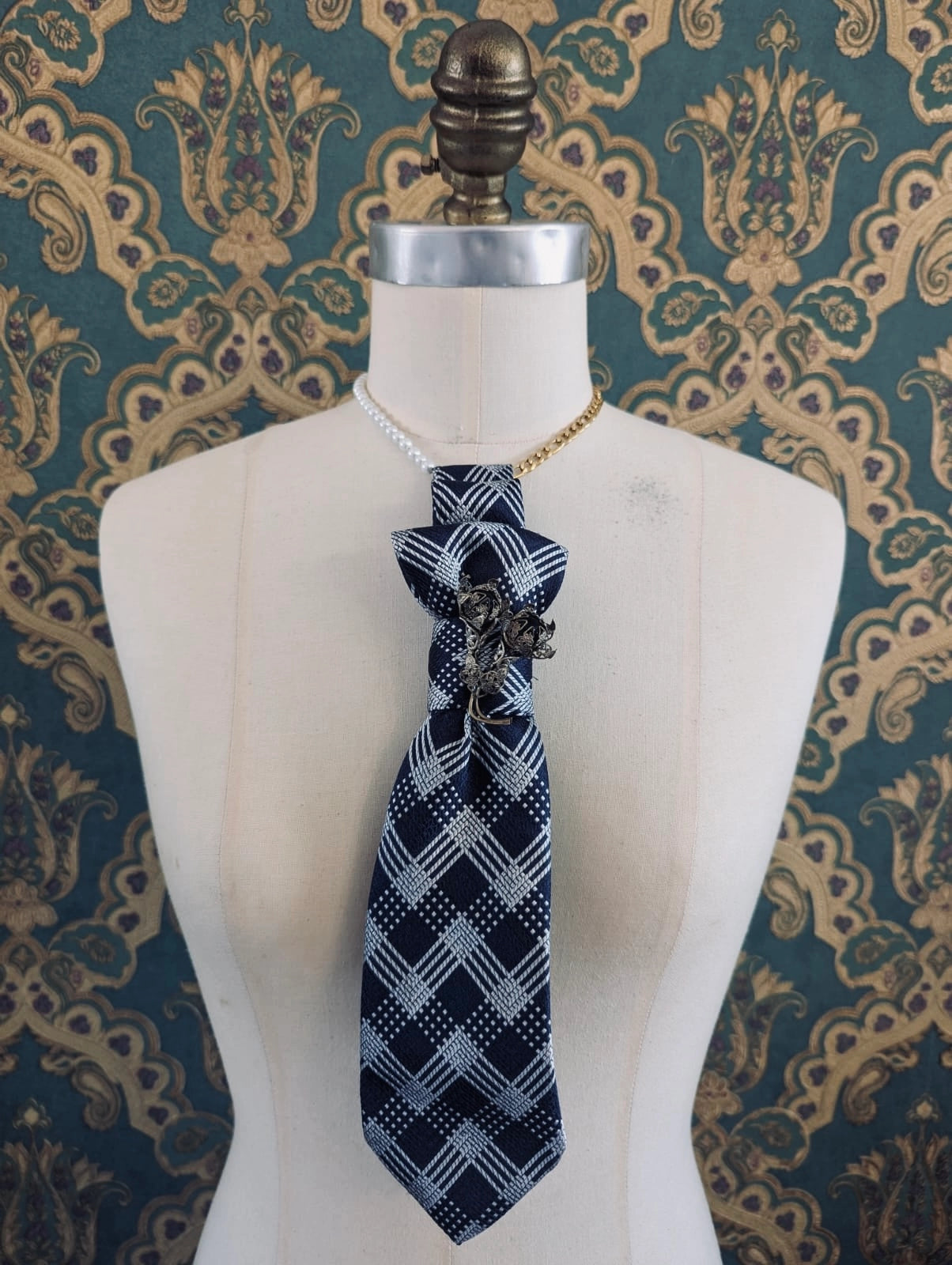 A Broochella montecristo cravatta navy blue tie brooch with a white geometric plaid pattern and a silver rose embellishment, displayed on a cream-colored dress form mannequin against a teal and gold damask patterned wallpaper background.