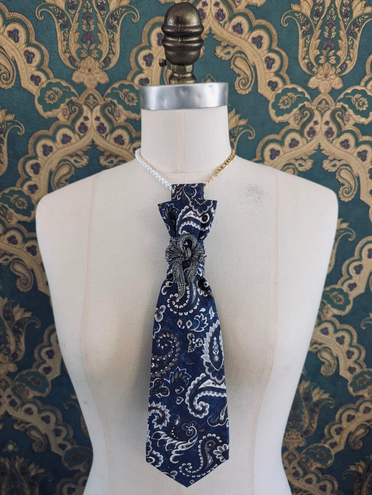 A Broochella Montecristo cravatta navy blue paisley handmade, upcycled tie brooch with white and silver detailing, styled on a cream-colored mannequin torso. The tie is accessorized with a pearl collar and a gold chain, set against a dark teal and gold patterned wallpaper background.
