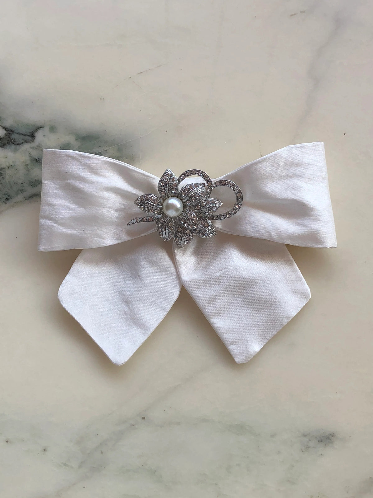 Broochella Moonlight Petal Bow Brooch in ivory silk featuring an ornate silver-tone floral embellishment with crystals and a central pearl, photographed on a pale white marble background with grey veining.