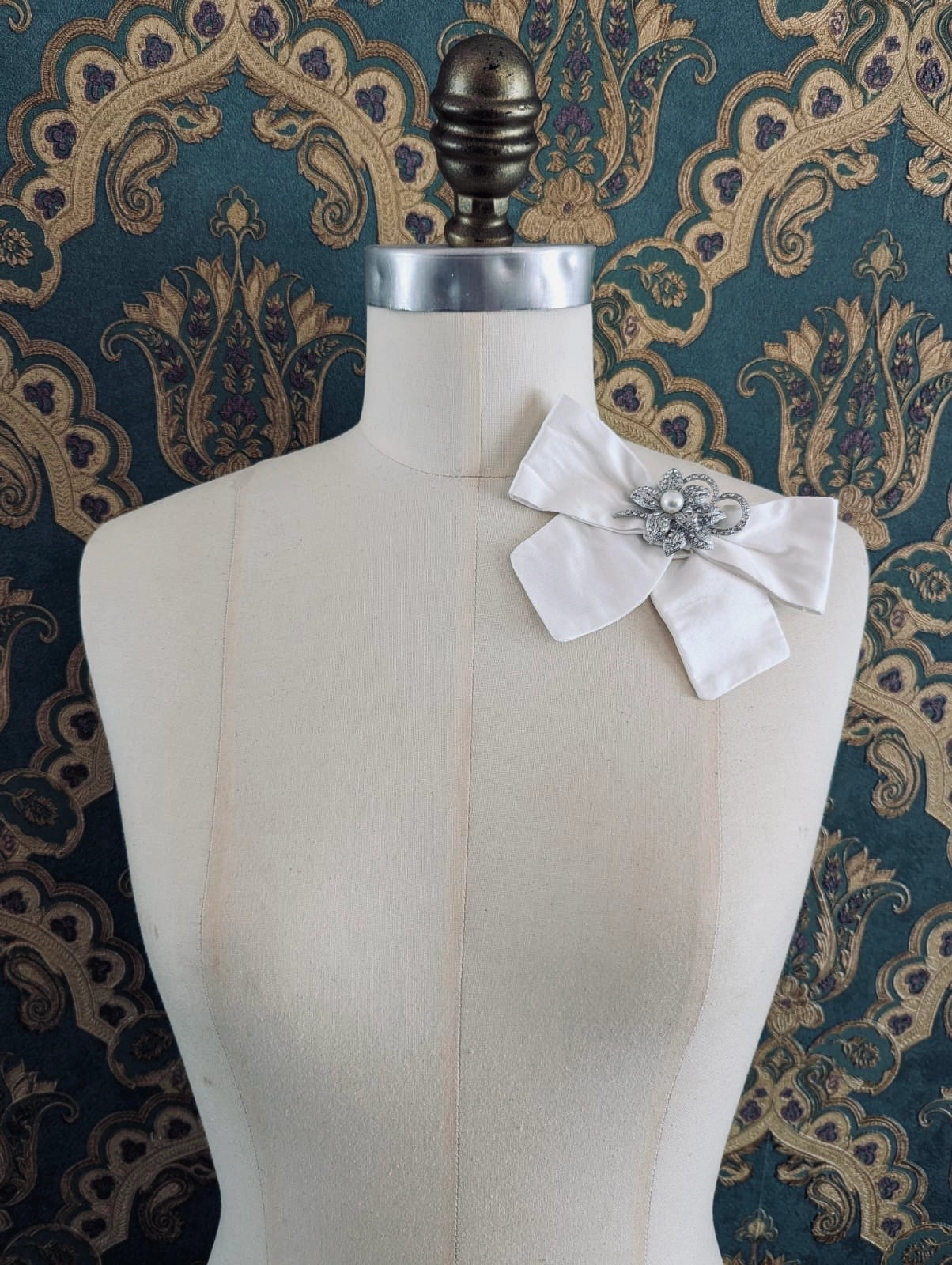 Broochella moonlight petal ivory silk bow brooch, with vintage brooch centre embellishment. Pinned on a mannequin. The background is green and gold damask wallpaper. 