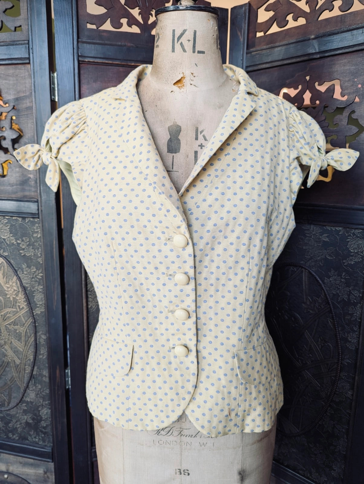 Broochella Moschino Cheap and Chic designer short sleeved jacket. It is pale yellow with blue polka dots. It is presented on a vintage mannequin. The background is an ornate, dark wood screen. 