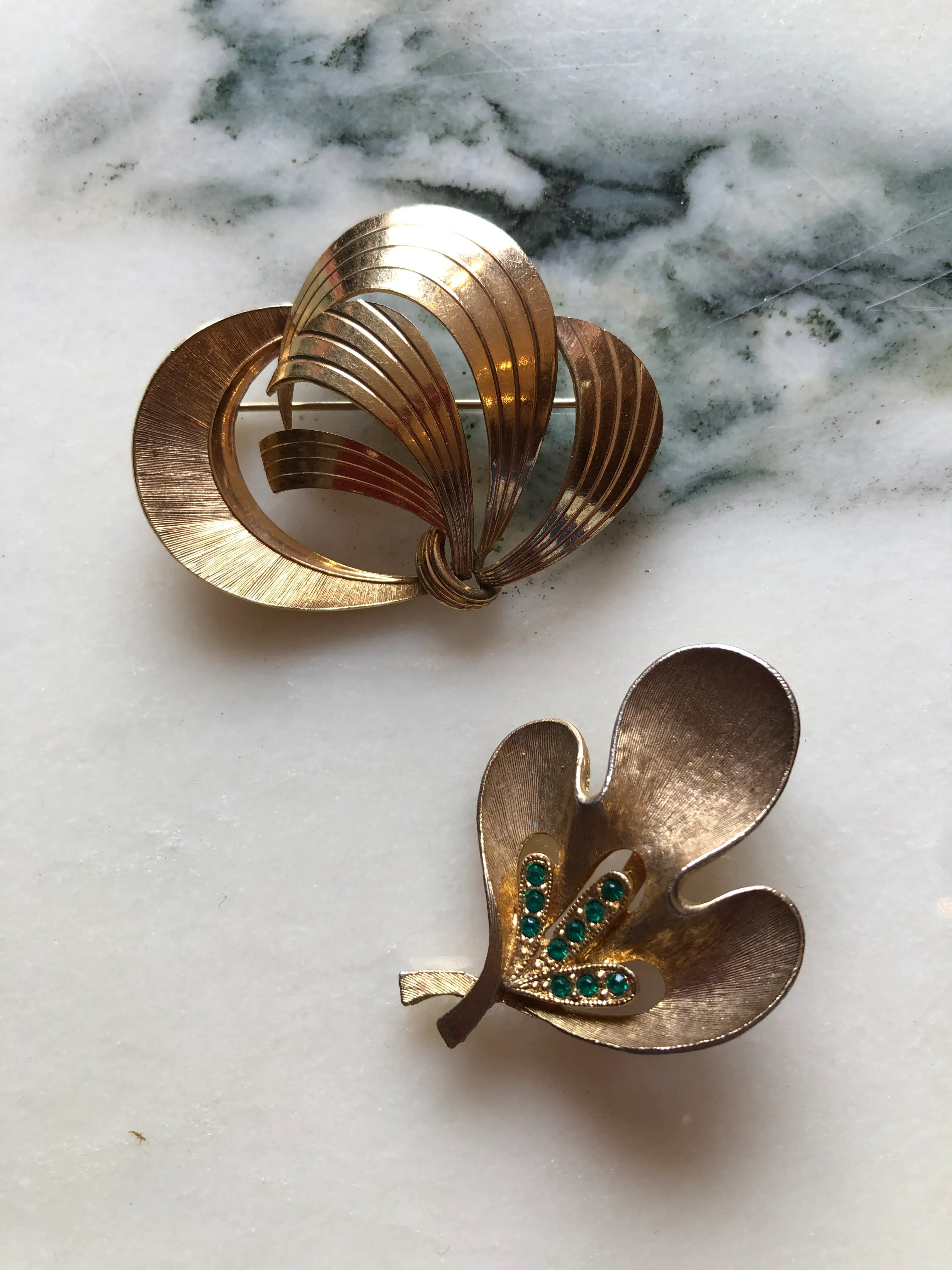 Broochella Movimento vintage modernist gold-tone brooch duo with an abstract ribbon brooch and a J.J. brooch featuring green crystals. The background is white marble