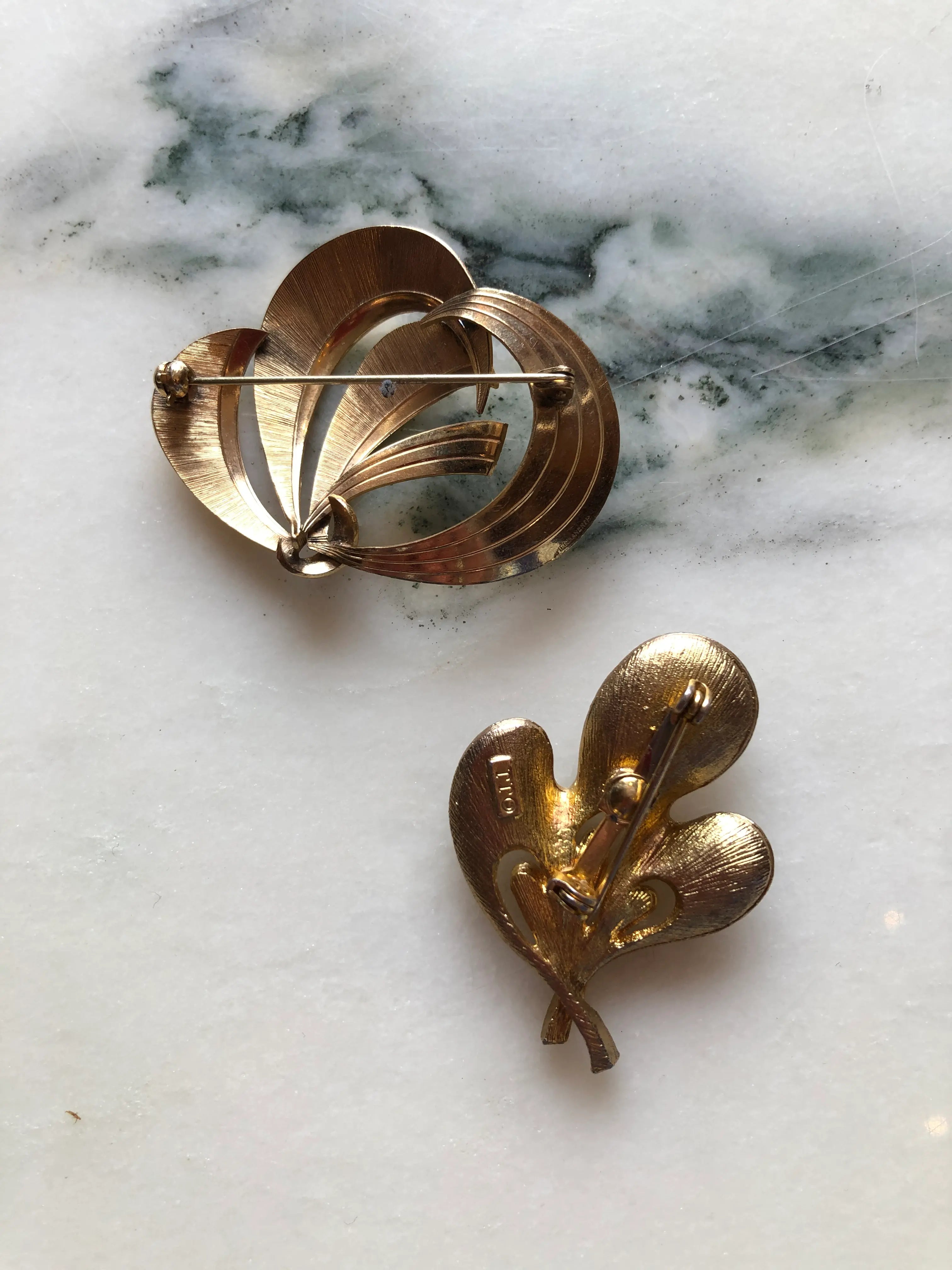 The back view of Broochella Movimento vintage modernist gold-tone brooch duo with an abstract ribbon brooch and a J.J. brooch featuring green crystals. The background is white marble