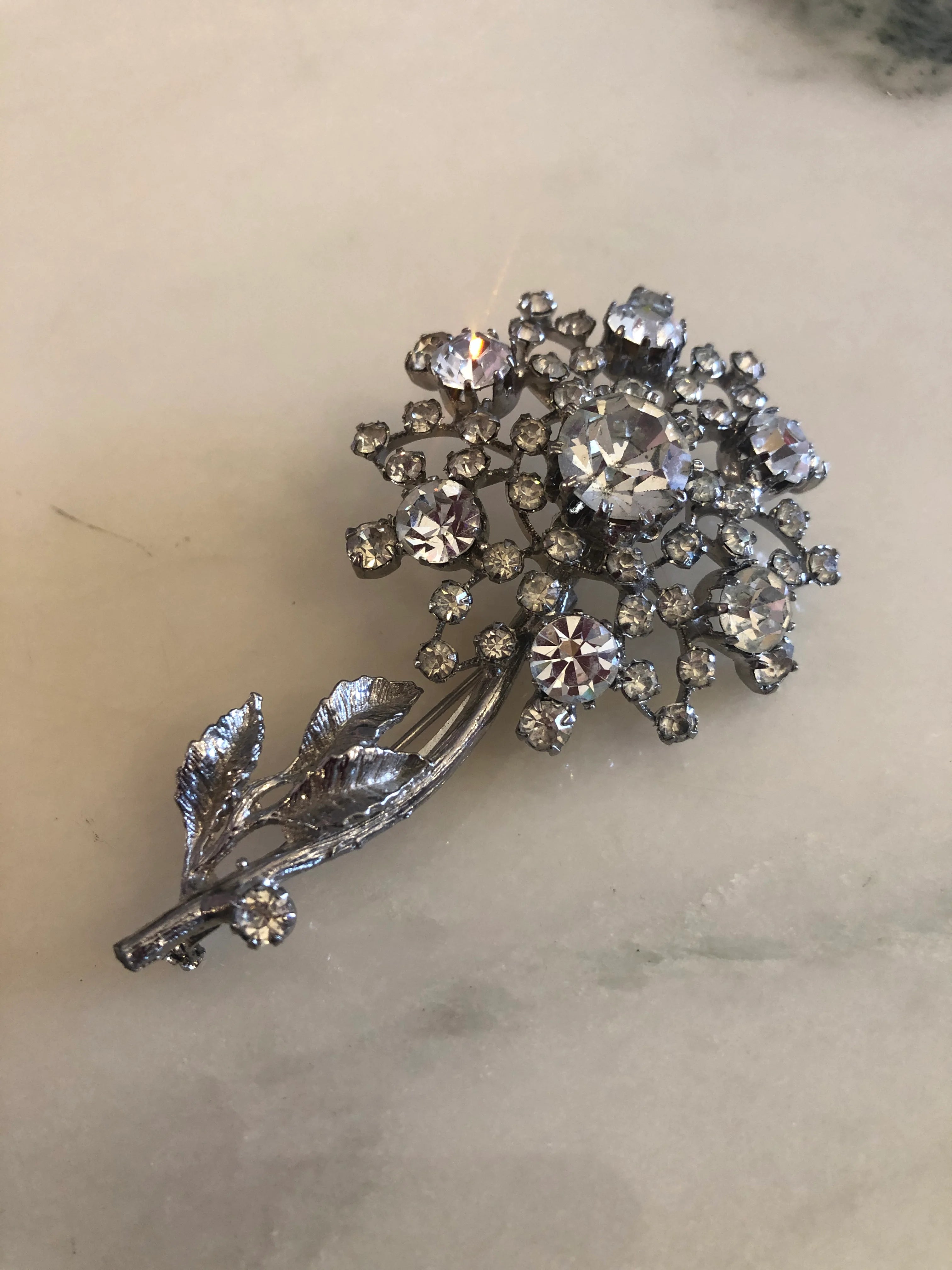 A Broochella Neve vintage flower brooch featuring a cluster of clear rhinestones and a leaf motif, displayed on white marble. 