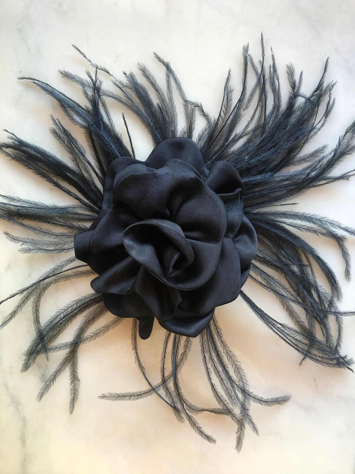Broochella Nocturne Black Flower & Feather Brooch featuring a voluminous black flower with softly folded sculptural petals, surrounded by long, wispy black feathers radiating outwards, photographed on pale white marble