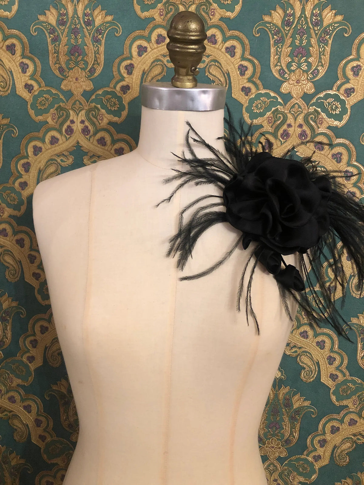 Broochella Nocturne Black Flower & Feather Brooch featuring an oversized sculptural black flower with voluminous layered petals and dramatic sprays of delicate black feathers, styled on the shoulder of an ivory mannequin against ornate emerald green and gold damask wallpaper.