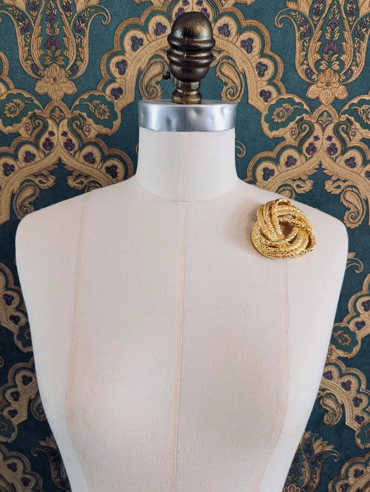 Broochella nodo golden knot metal designer brooch. Pictured on a mannequin. The background is green and gold damask wallpaper