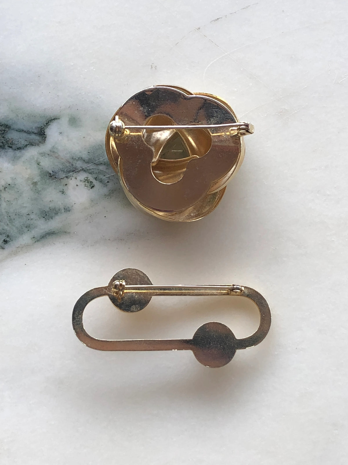 Broochella Noeud and Perle Vintage Brooch Duo back view showing the gold-tone settings and pin fastenings of both vintage brooches, photographed on a white marble background with soft grey-green veining.