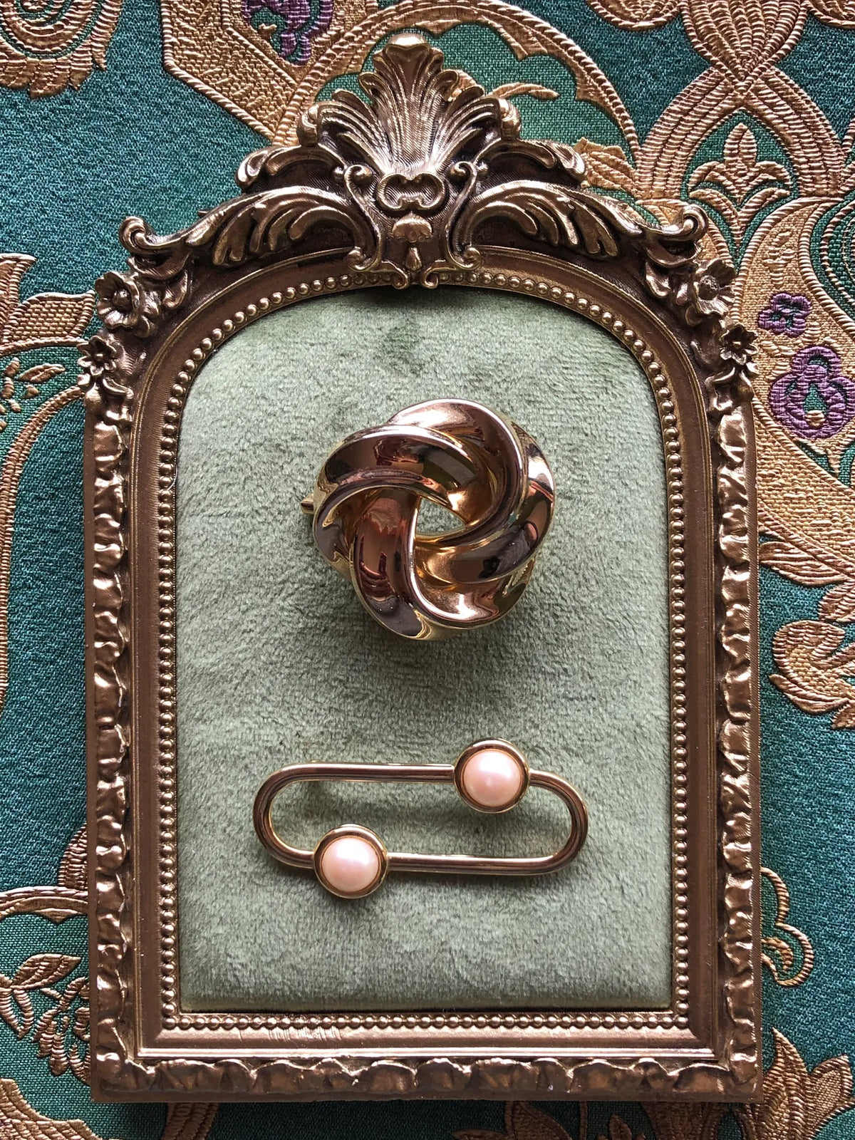 Broochella Noeud and Perle Vintage Brooch Duo featuring a polished gold-tone interwoven knot brooch and an oval gold-tone brooch with two luminous faux pearls, displayed on sage green velvet in an ornate antique gold frame against green and gold patterned fabric.