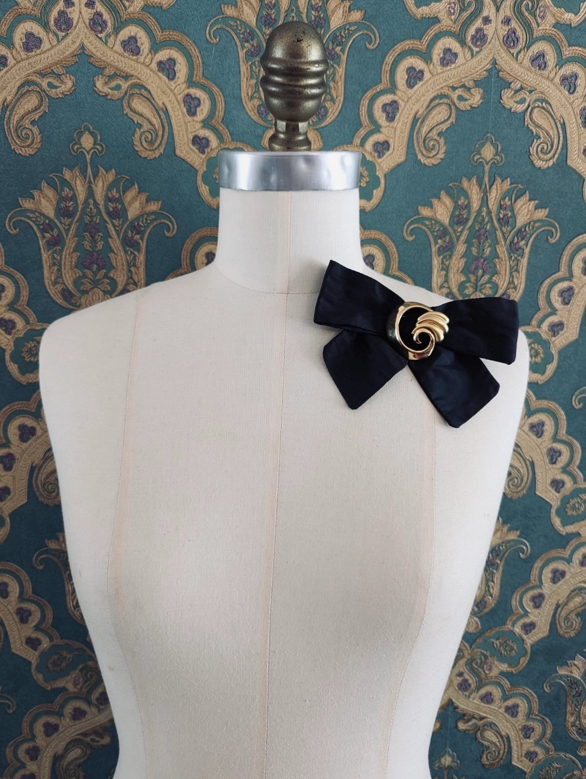 Broochella noir decadence handmade designer silk bow brooch, with gold vintage brooch embellishment. Pinned to a mannequin. The background is green and gold smamsk wallpaper
