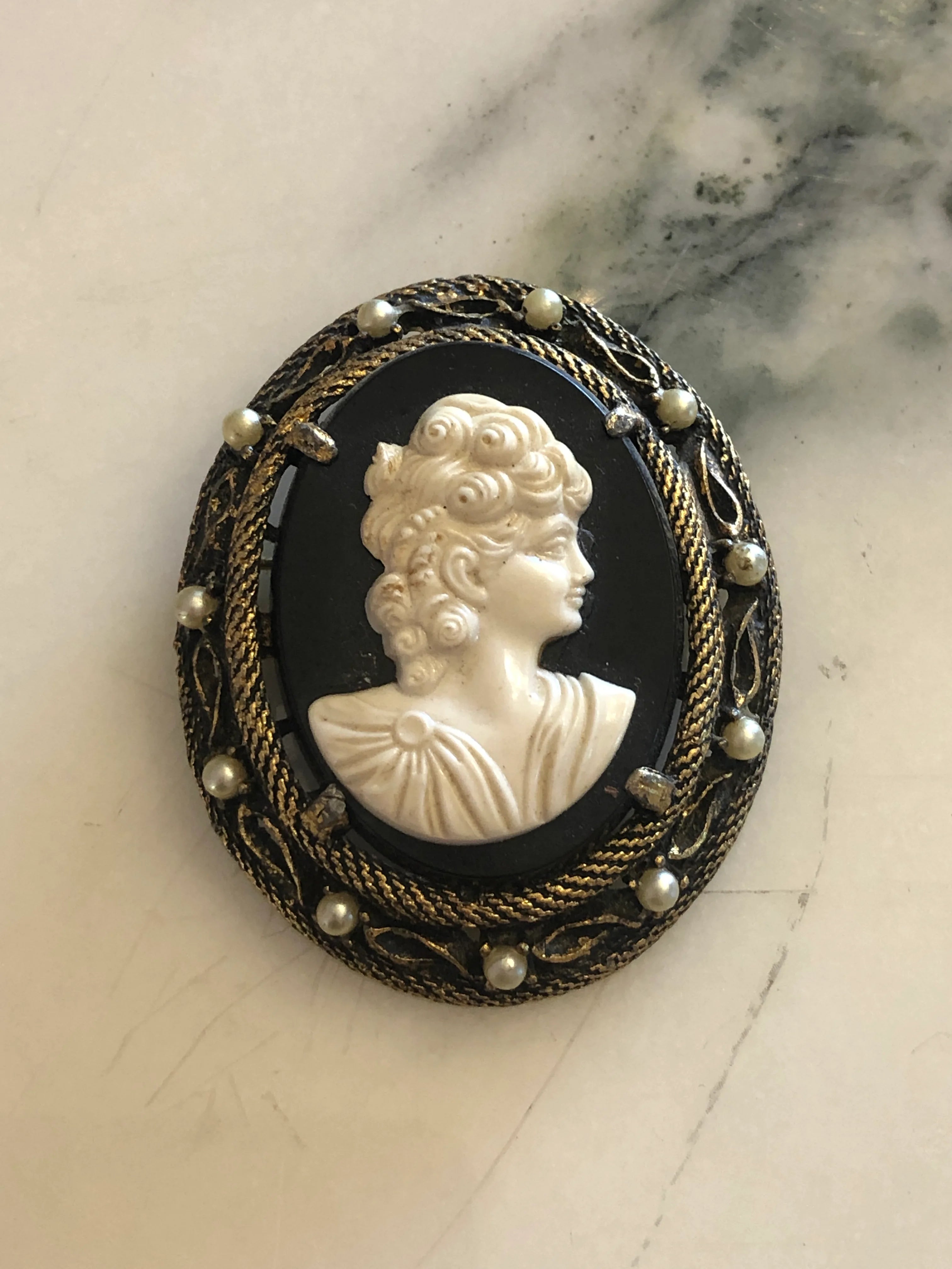 A 5.5cm x 4.5cm vintage cameo brooch featuring a carved white classical portrait on a deep black ground set within a gold-tone rope twist filigree frame with seed pearl accents, photographed on a white marble surface.