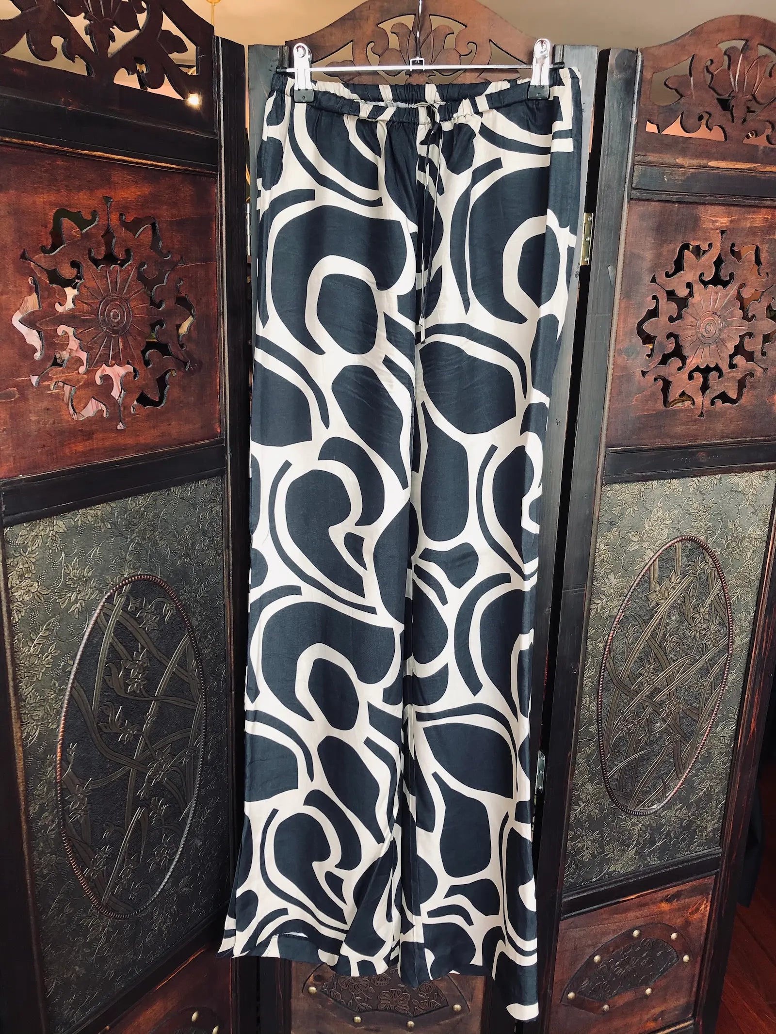 Broochella The Stylist's Closet Ode  wide-leg pants with an abstract grey and off-white swirl pattern, hanging on a wooden hanger against a dark carved wooden folding screen.