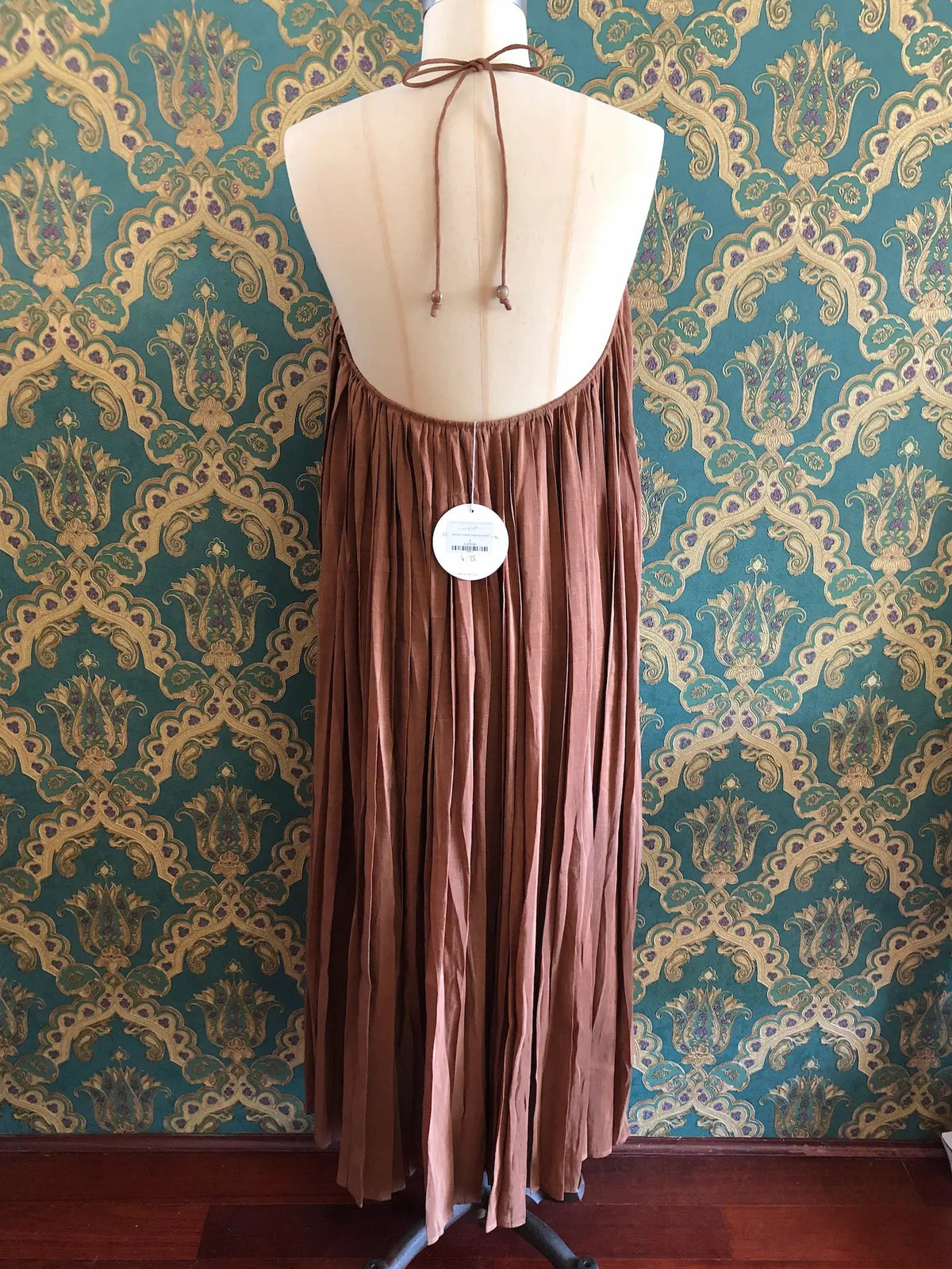 Odet Tropicale cocoa linen and silk sunray-pleated sundress with beaded halter straps, size Small. Pictured on a mannequin. The background is green and gold damask wallpaper.