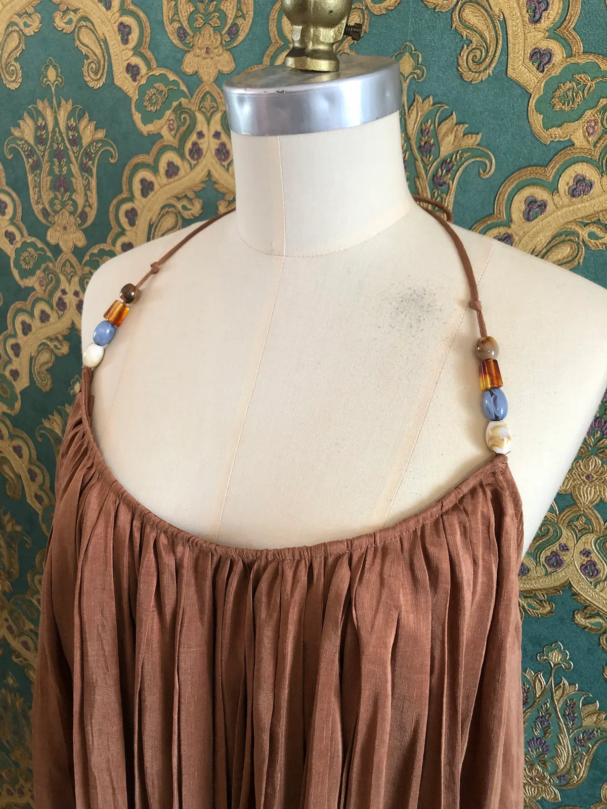 Odet Tropicale cocoa linen and silk sunray-pleated sundress with beaded halter straps, size Small. Pictured on a mannequin. The background is green and gold damask wallpaper.