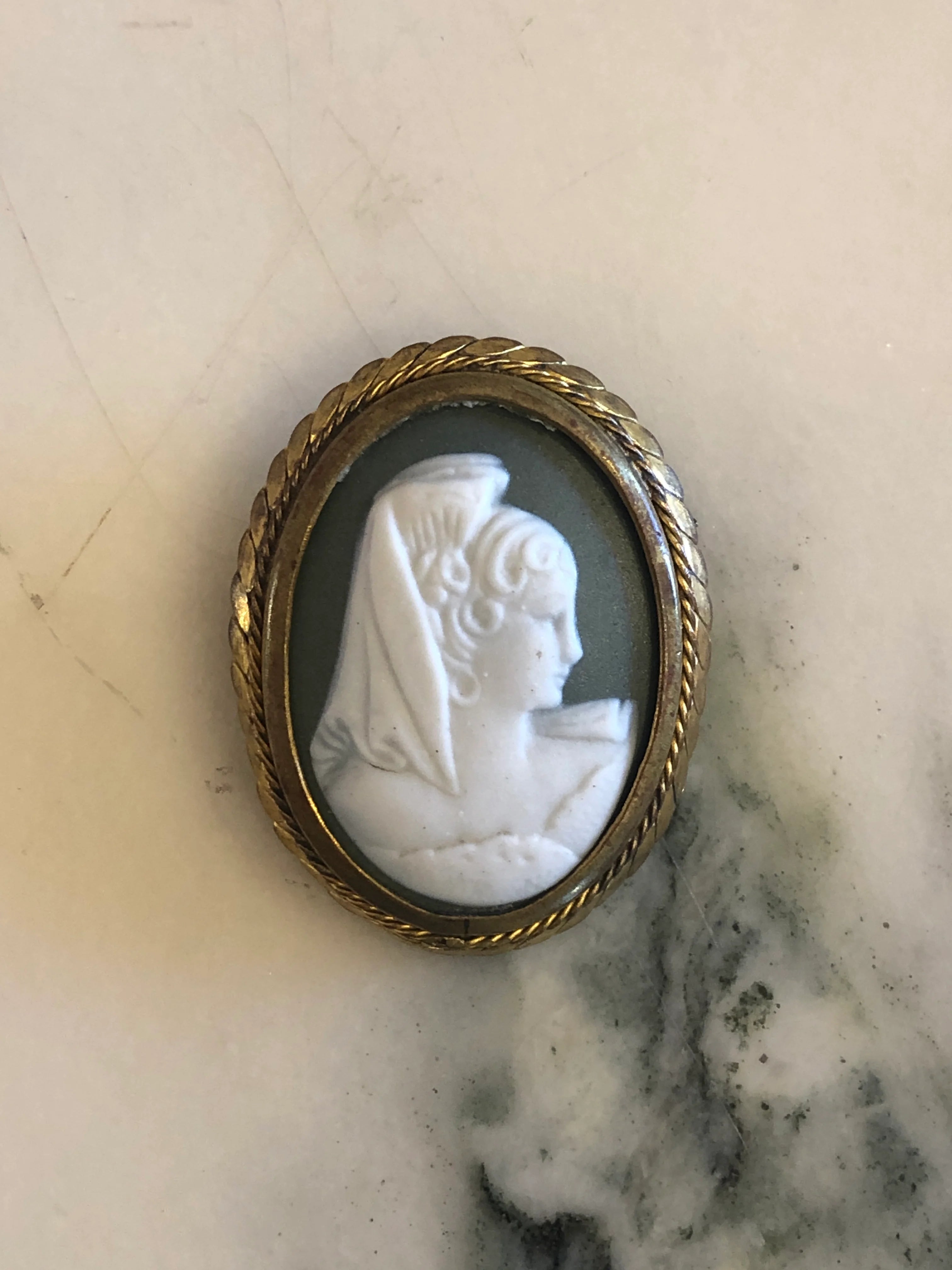Broochella Ombra vintage cameo brooch in green and white, with a gold tone metal frame. The background is white marble. 
