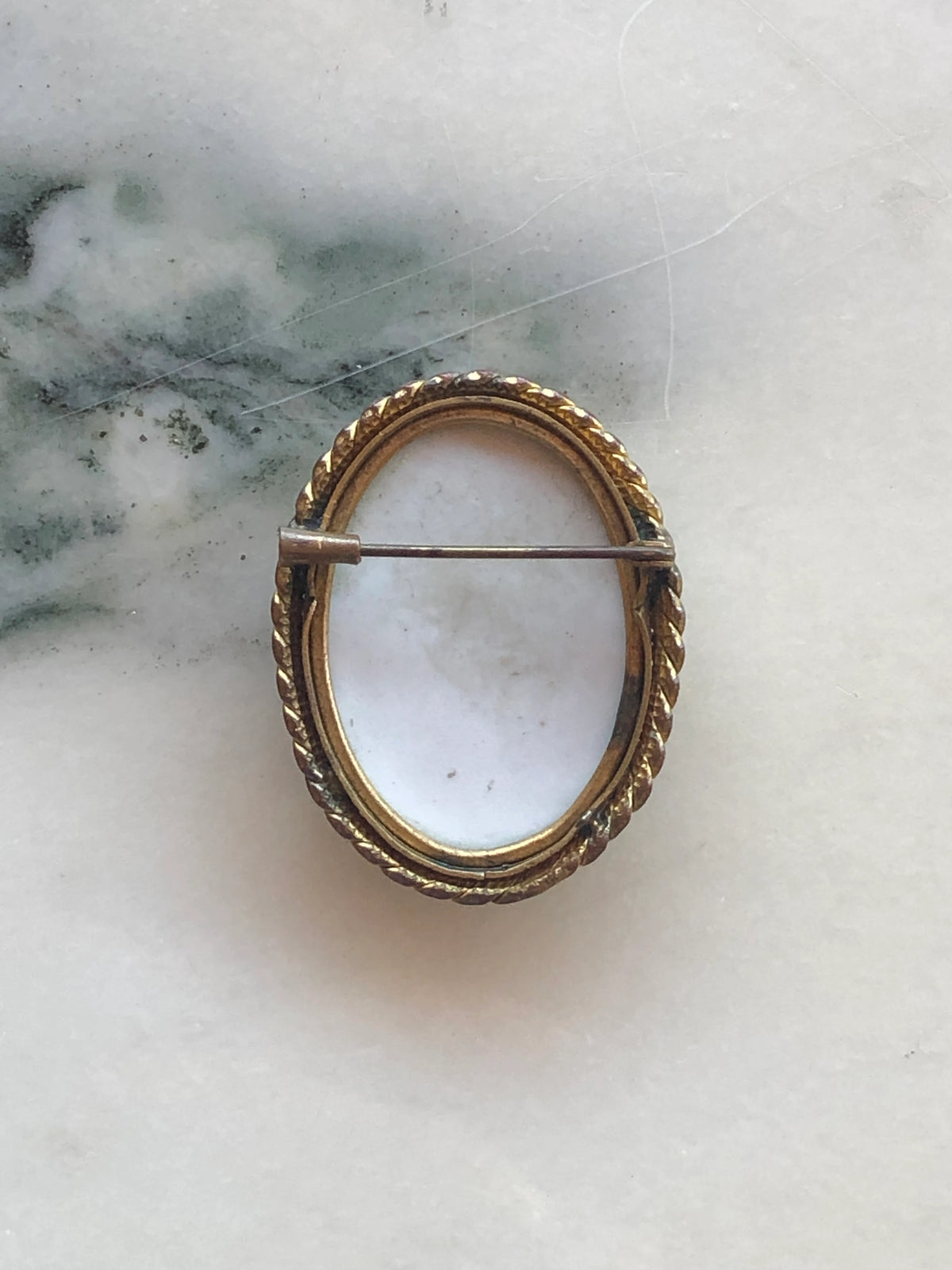 The back view of Broochella Ombra vintage cameo brooch in green and white, with a gold tone metal frame. The background is white marble. 