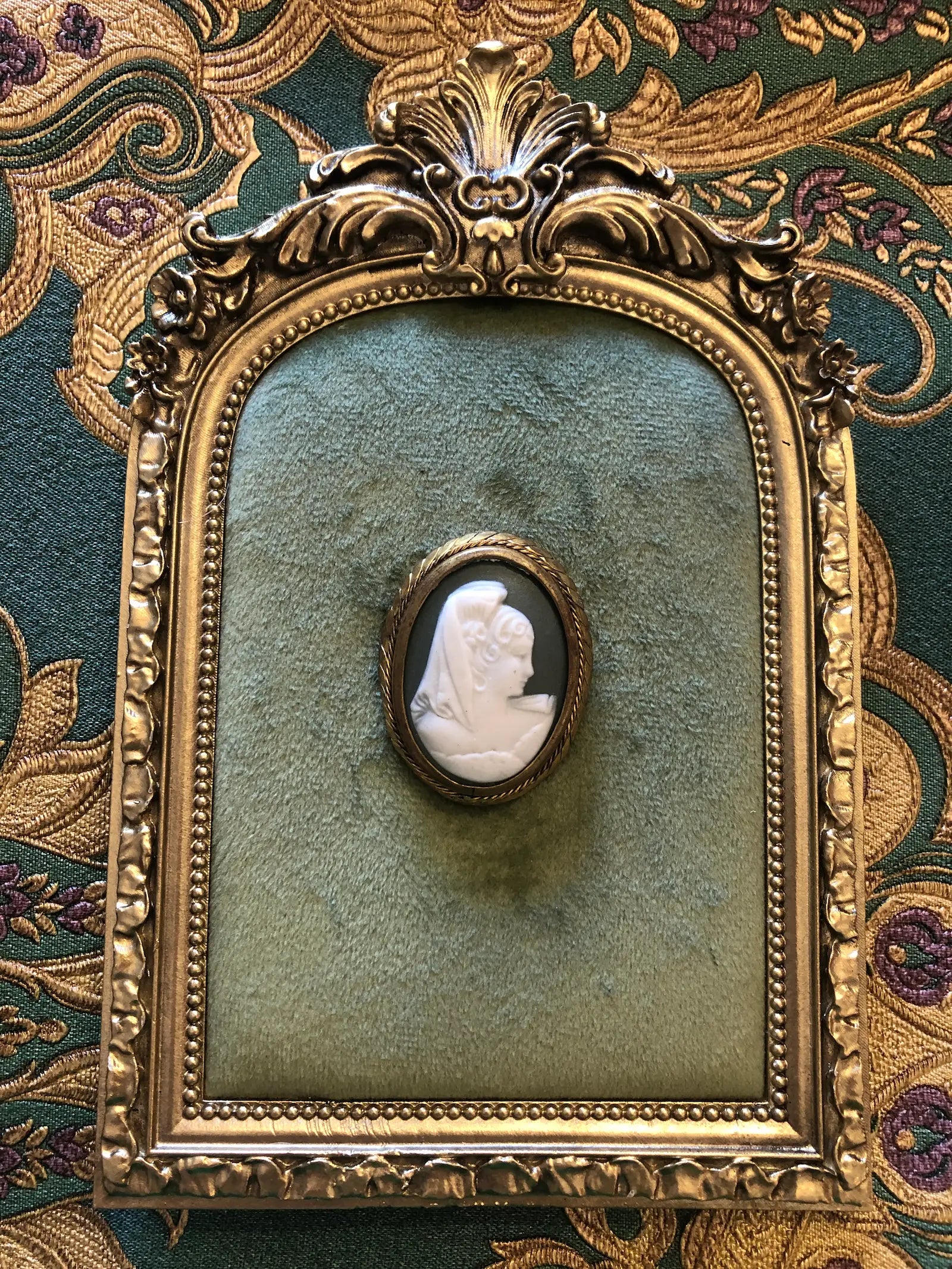 An ornate vintage-style gold frame with a green velvet center, featuring a  Broochella ombra vintage cameo brooch in the middle, set against a patterned green and gold fabric background.