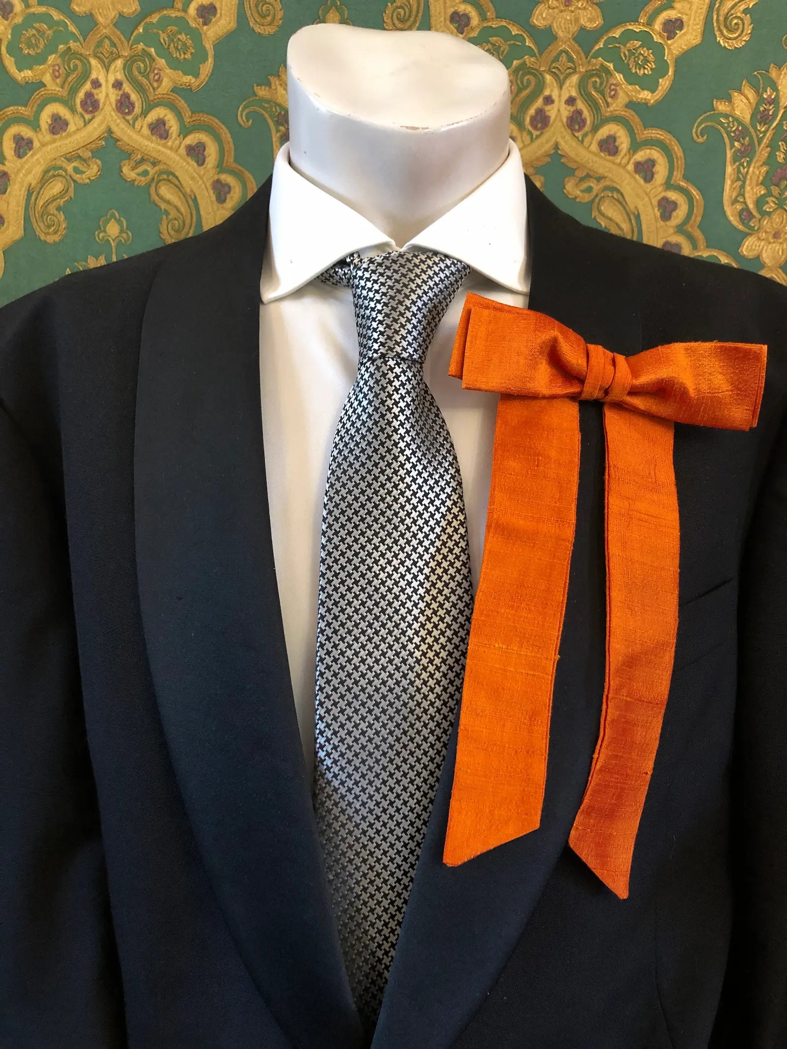 A Broochella Men's Alice silk bow brooch in orange. It is styled on a white shirt and patterned tie, displayed on a black suit jacket mannequin torso against a green and gold damask wallpaper background.