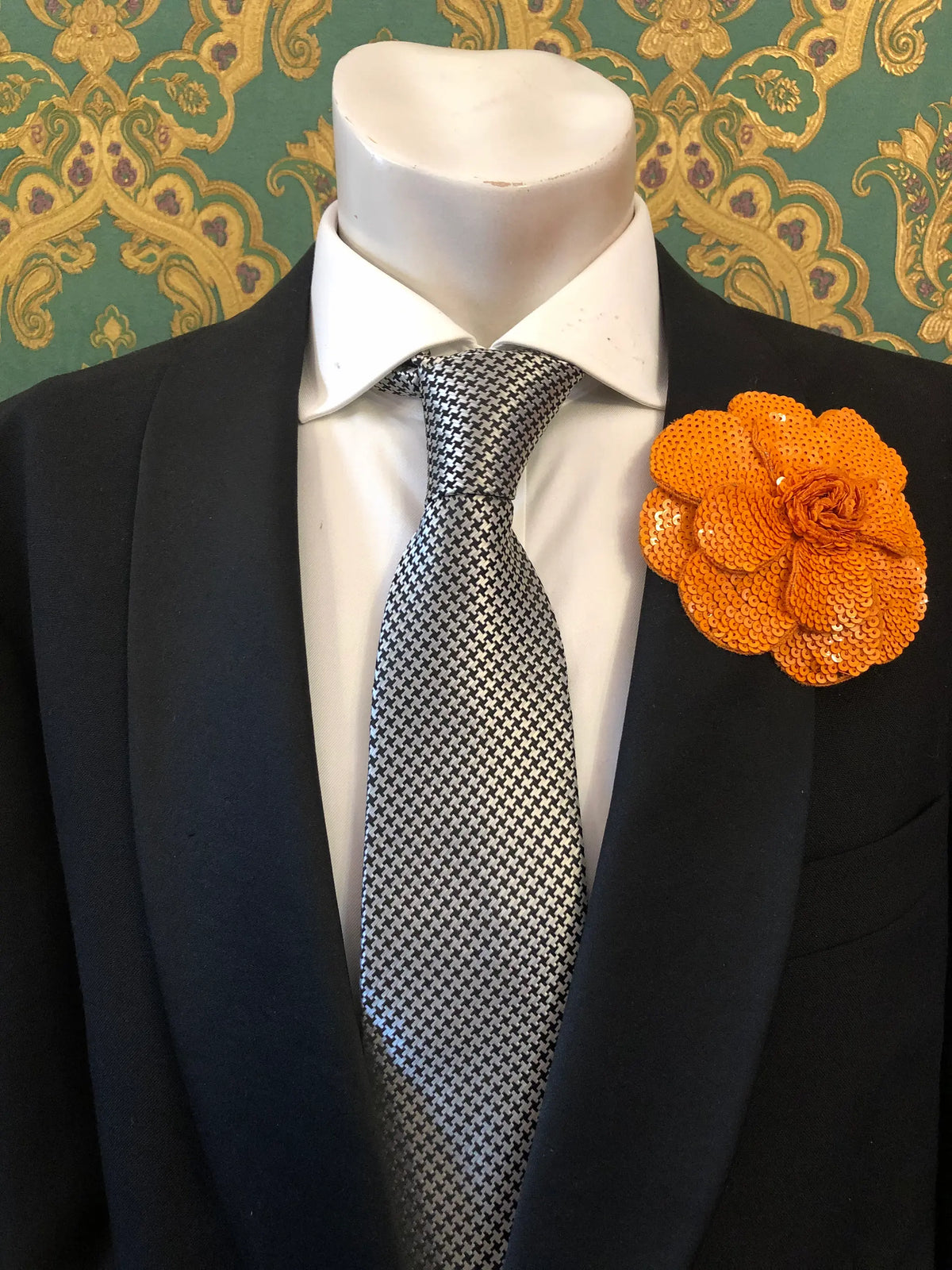 A men's formal accessory set displayed on a white mannequin torso, featuring a black suit jacket, a white dress shirt, a silver and black checkered tie, and an orange Men's Broochella Scintillante sequinned flower brooch lapel pin against a green and gold patterned wallpaper background.