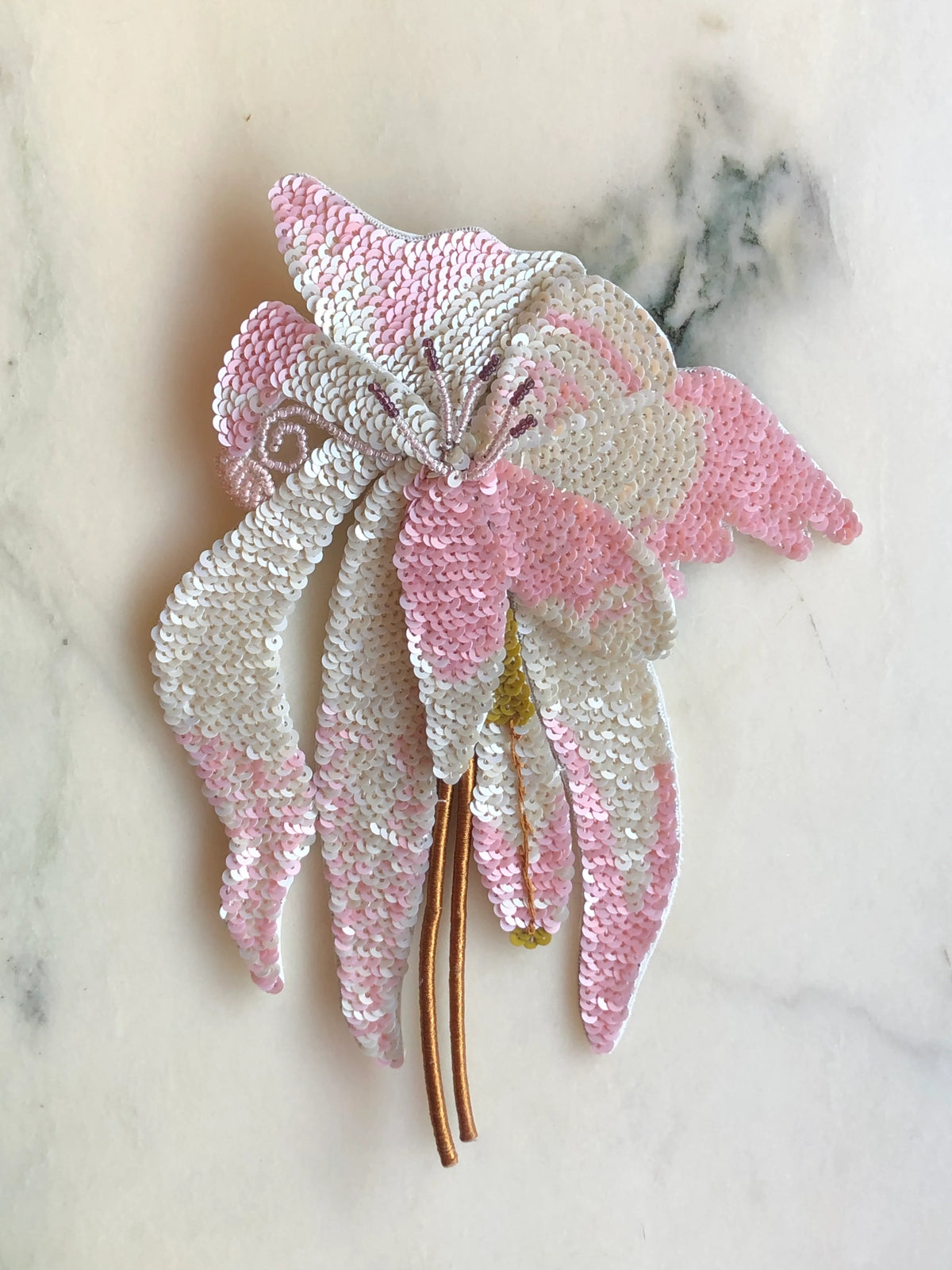 Broochella Men's Orchid Sequin Flower Brooch Pin embellished with soft pink and white sequins, featuring sculptural orchid petals, delicate beaded details and long gold stems, photographed on a white marble background with soft grey-green veining.