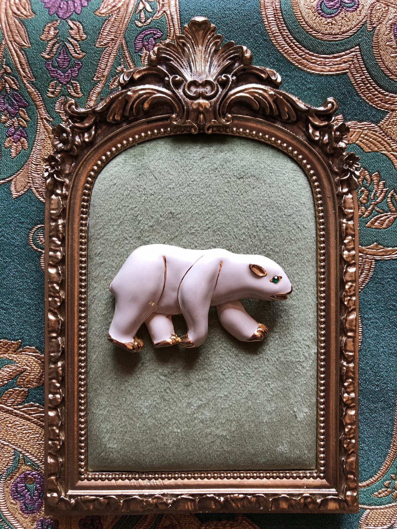 Broochella Orso Bianco vintage white enamel polar bear brooch with gold-tone details and a green crystal eye. Pictured on a green velvet tray with an ornate gold frame. The background is green and gold damask wallpaper. 