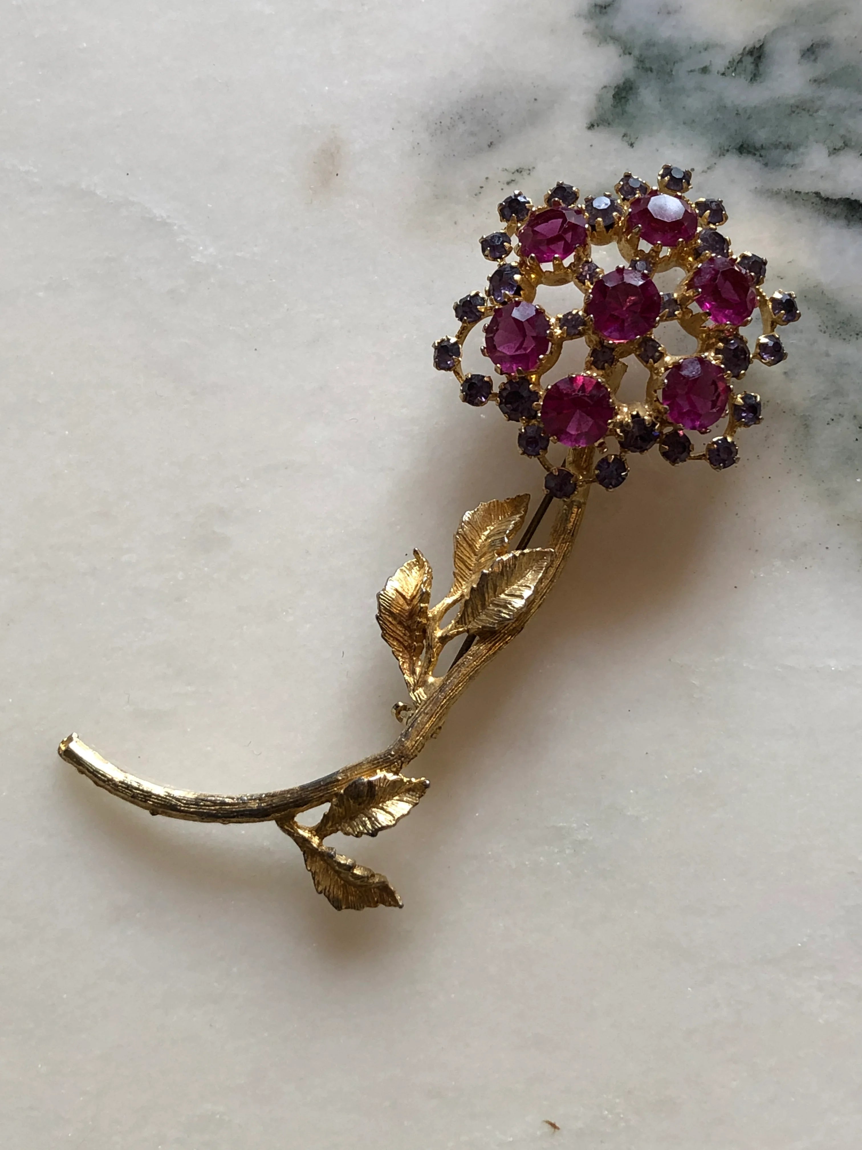 Broochella Ortensia Reale vintage gold-tone flower brooch with a long stem, textured leaves and fuchsia and purple crystals. The background is white marble