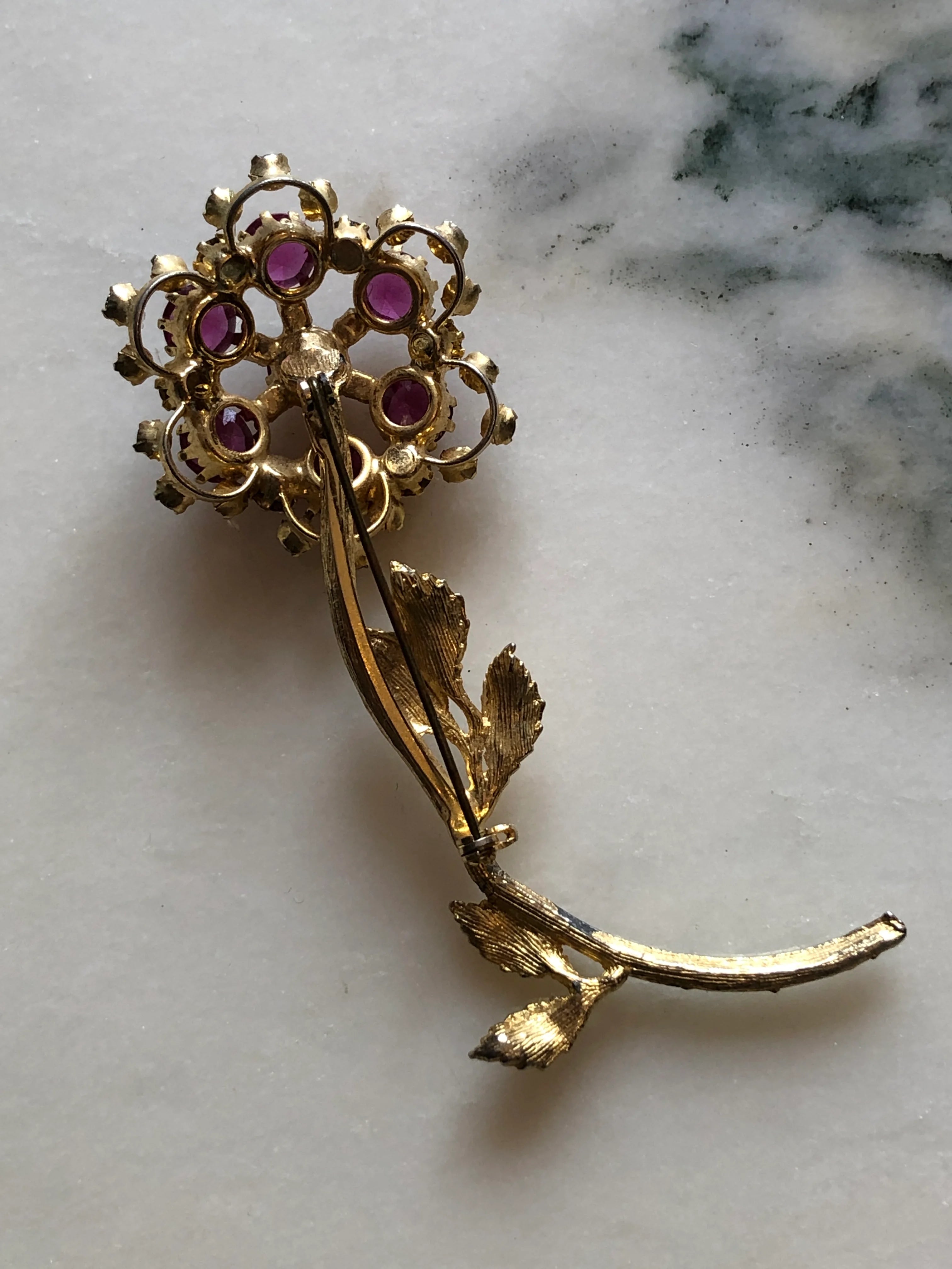 The back view of Broochella Ortensia Reale vintage gold-tone flower brooch with a long stem, textured leaves and fuchsia and purple crystals. The background is white marble. 
