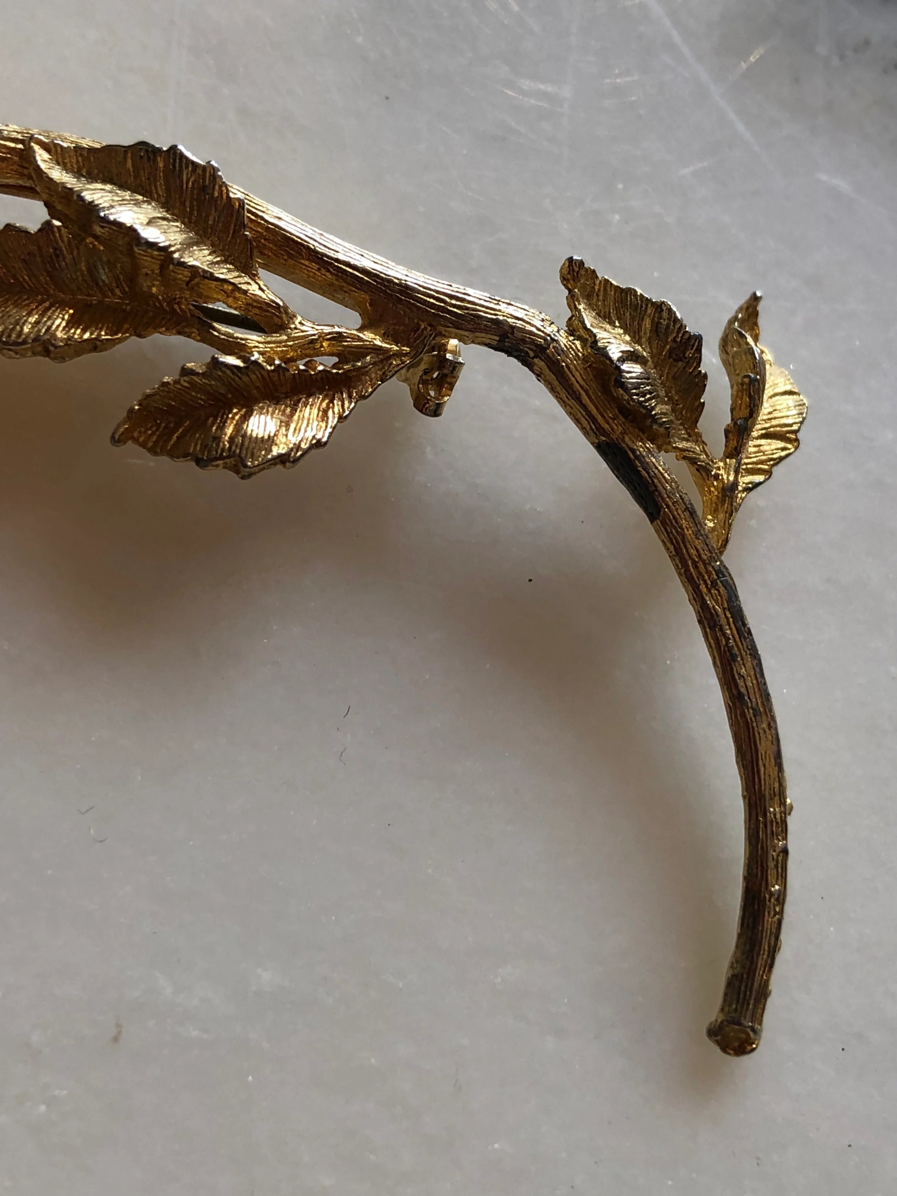 A close up of the stem of the Broochella Ortensia Reale vintage gold-tone flower brooch with a long stem, textured leaves and fuchsia and purple crystals. The background is white marble