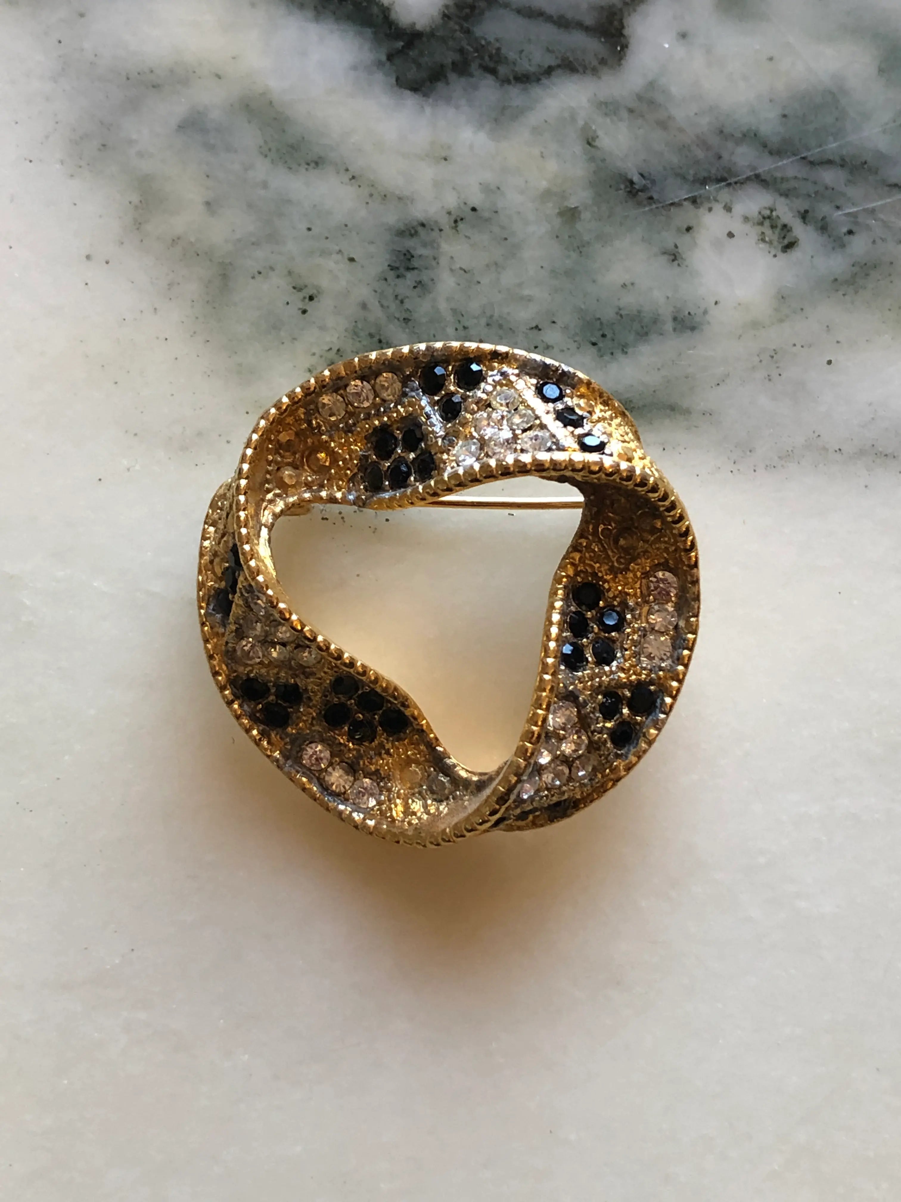 Broochella Ottavia Vintage Black & Crystal Swirl Brooch featuring black and clear crystals set into a sculptural gold-tone circular design on a cream and grey marble background.