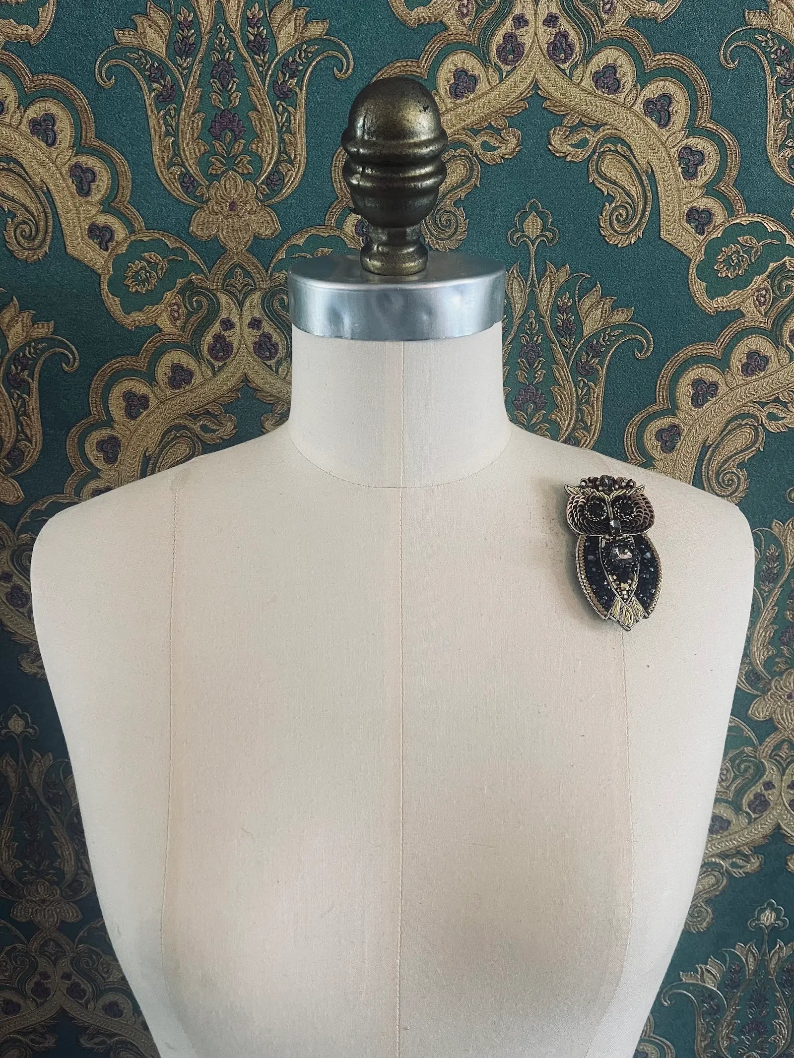 Broochella hand beaded gold and black owl brooch. Pictured on a mannequin. The background is green and gold damask wallpaper