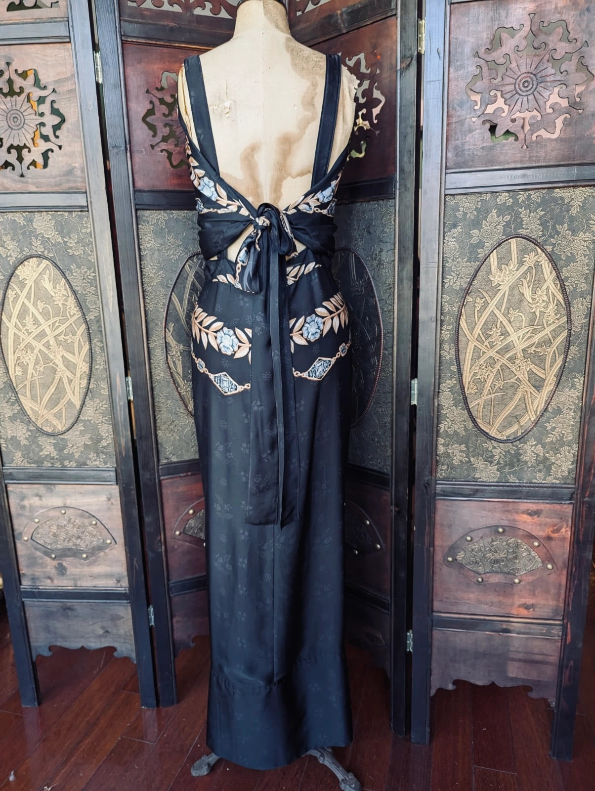 The back view of  a Broochella Paco Rabanne authentic designer black satin maxi dress with gem and flower print. Decorative black evening gown on a mannequin against an ornate wooden wall.