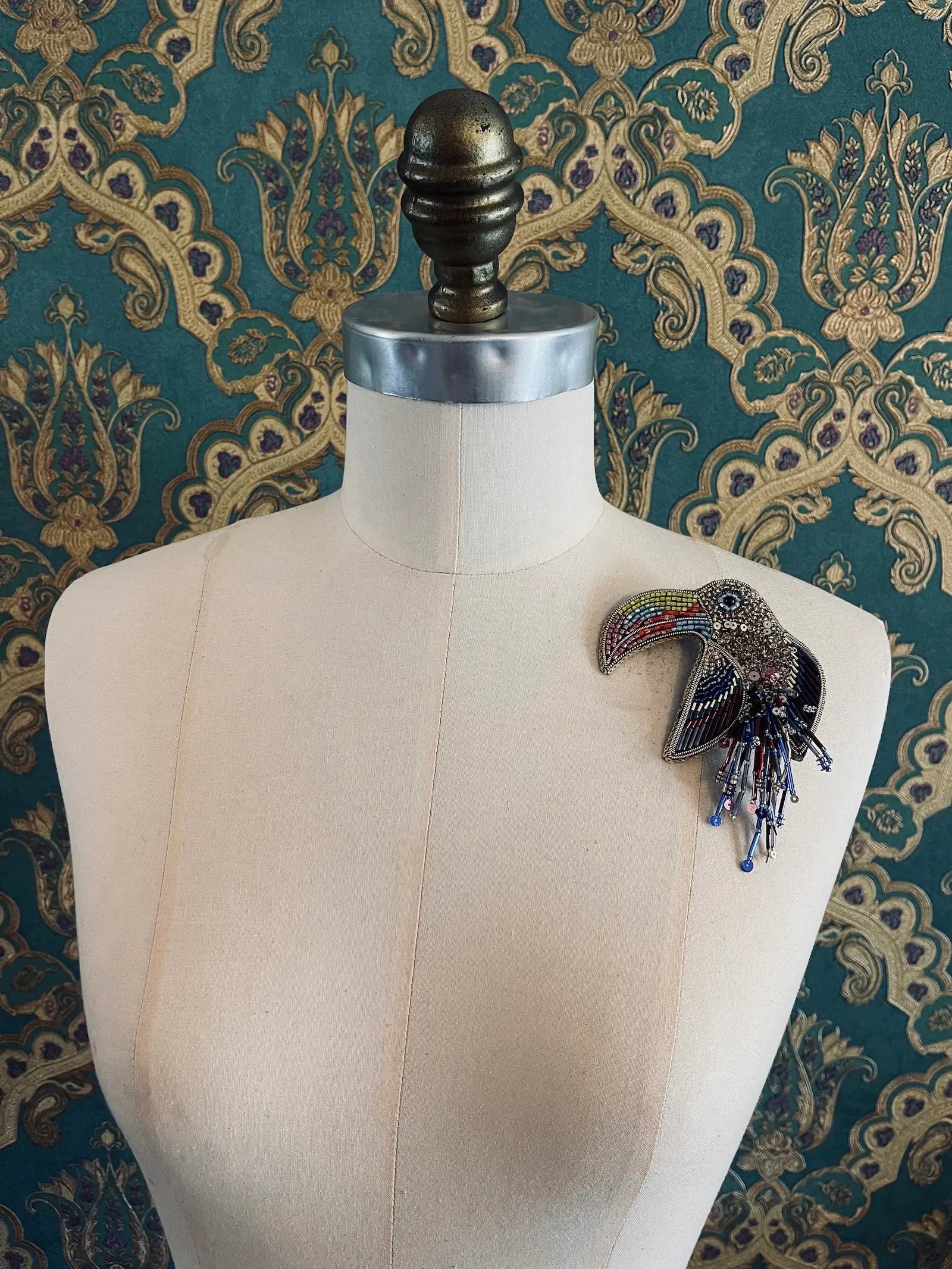 Broochella pappagallo multi coloured parrot bird brooch hand made from  beads and sequins. Pictured on a mannequin. The background is green and gold damask wallpaper.