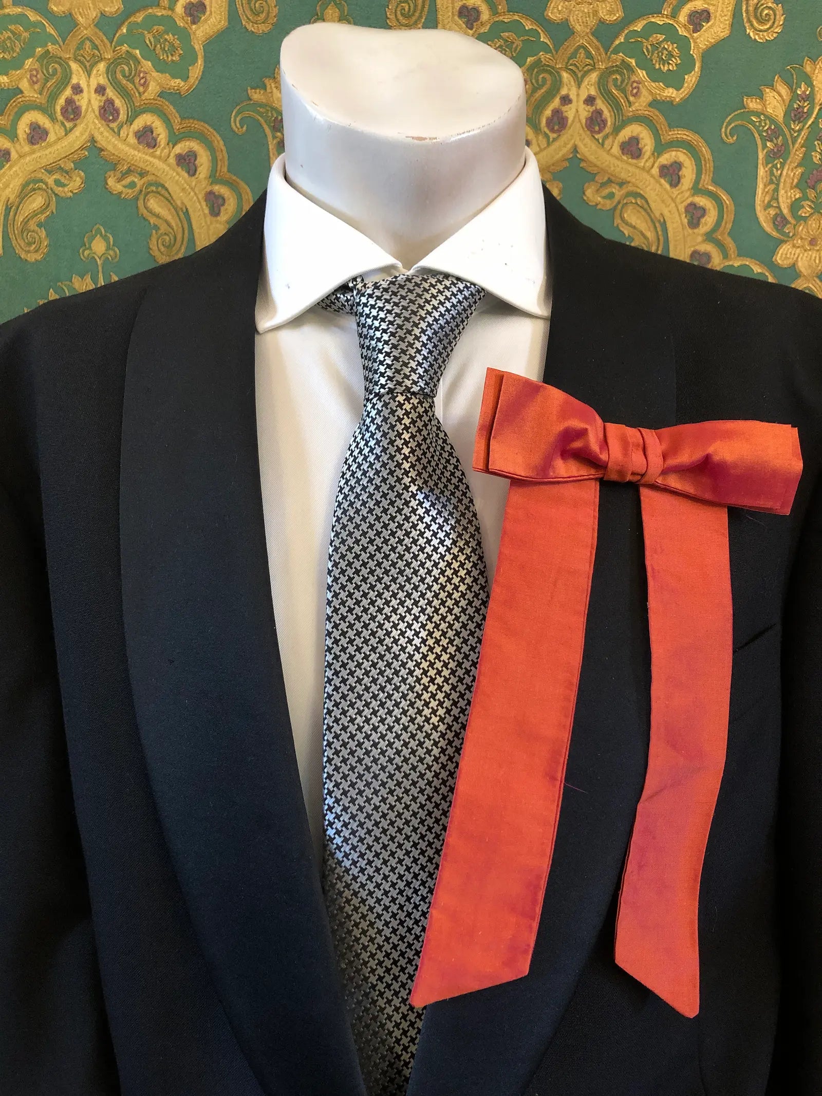 A Broochella Men's Alice silk bow brooch in peach. It is styled on a white shirt and patterned tie, displayed on a black suit jacket mannequin torso against a green and gold damask wallpaper background.