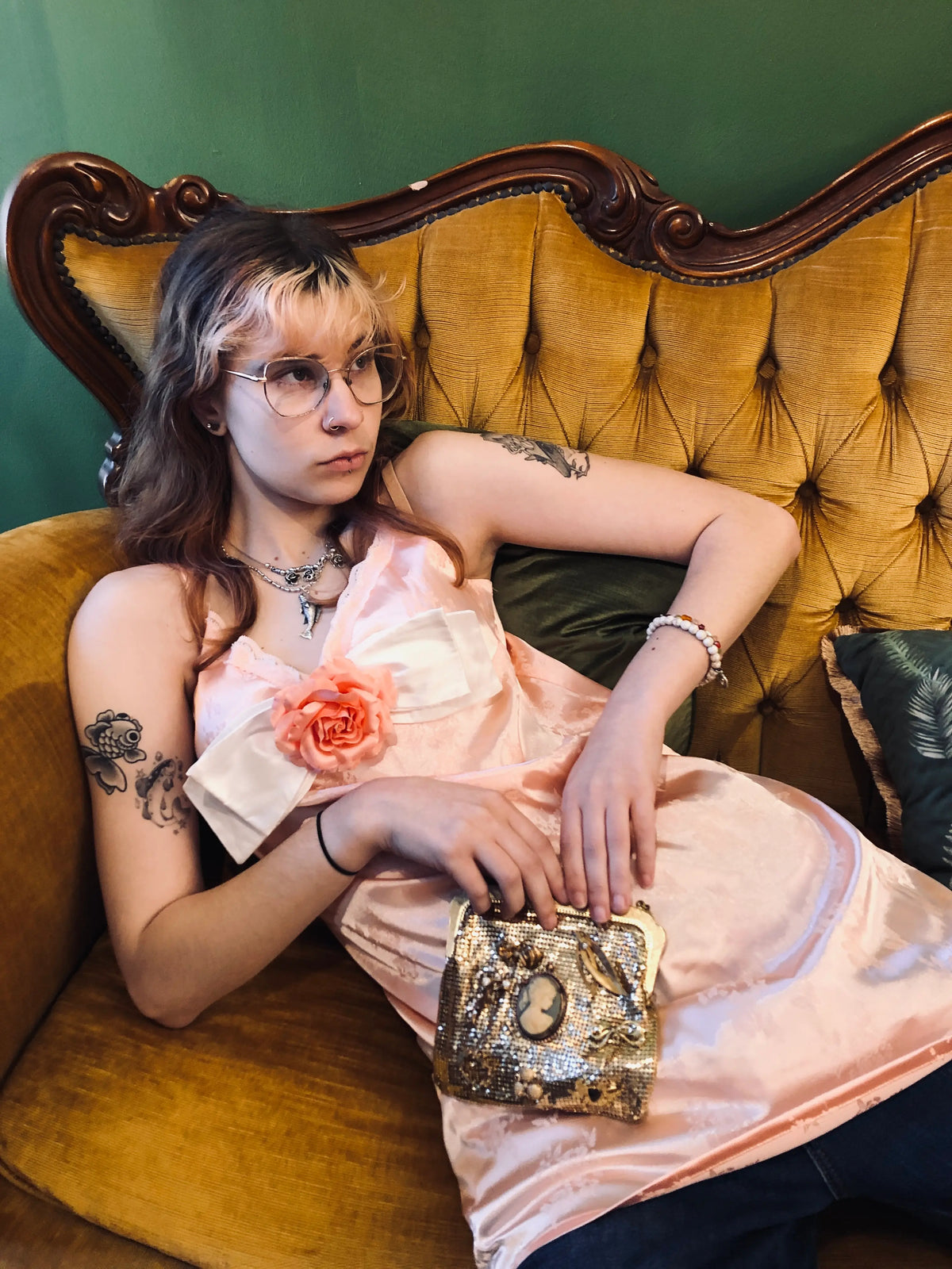 A woman with glasses and tattoos reclines on an ornate mustard-yellow tufted sofa, wearing a pale pink satin dress with a Broochella peaches and cream bow brooch with an ivory bow and  peach rose detail on the chest. She is holding a small metallic clutch. 