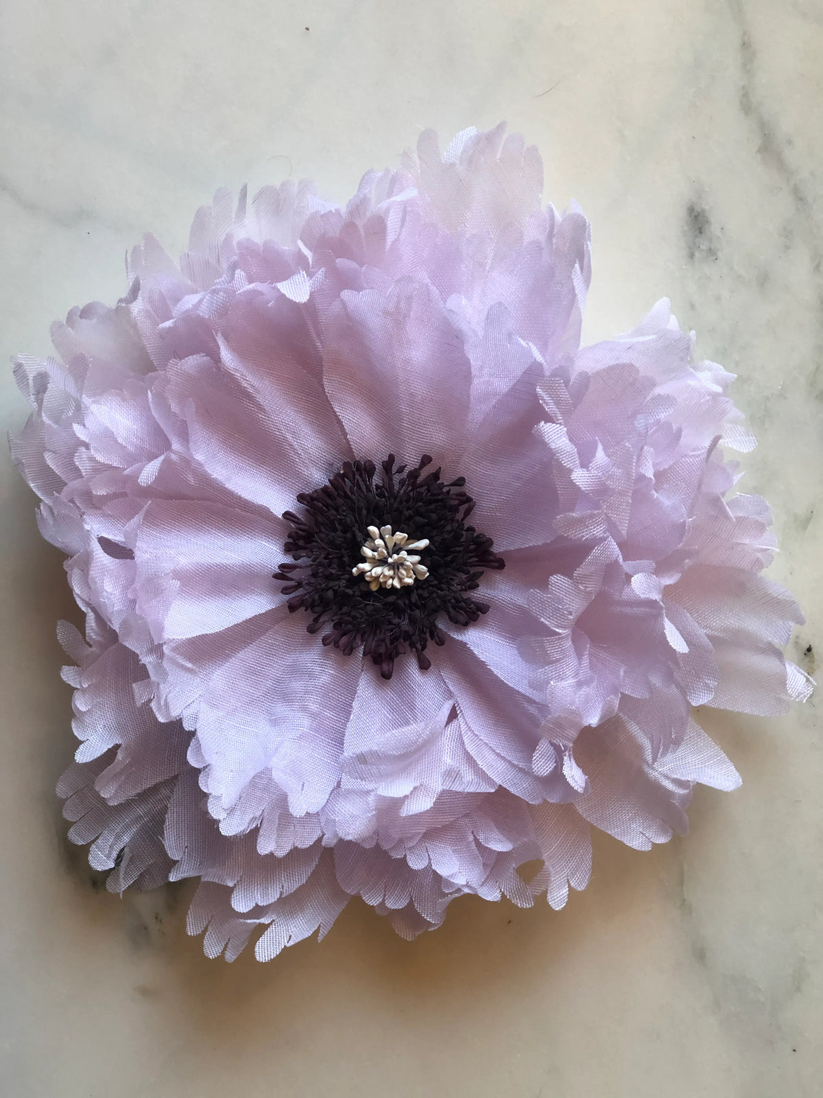 Broochella Peone Bridal Brooch Pin featuring layers of soft pale lilac fabric petals with a deep plum floral centre and delicate ivory detail, photographed on a pale cream marble background.