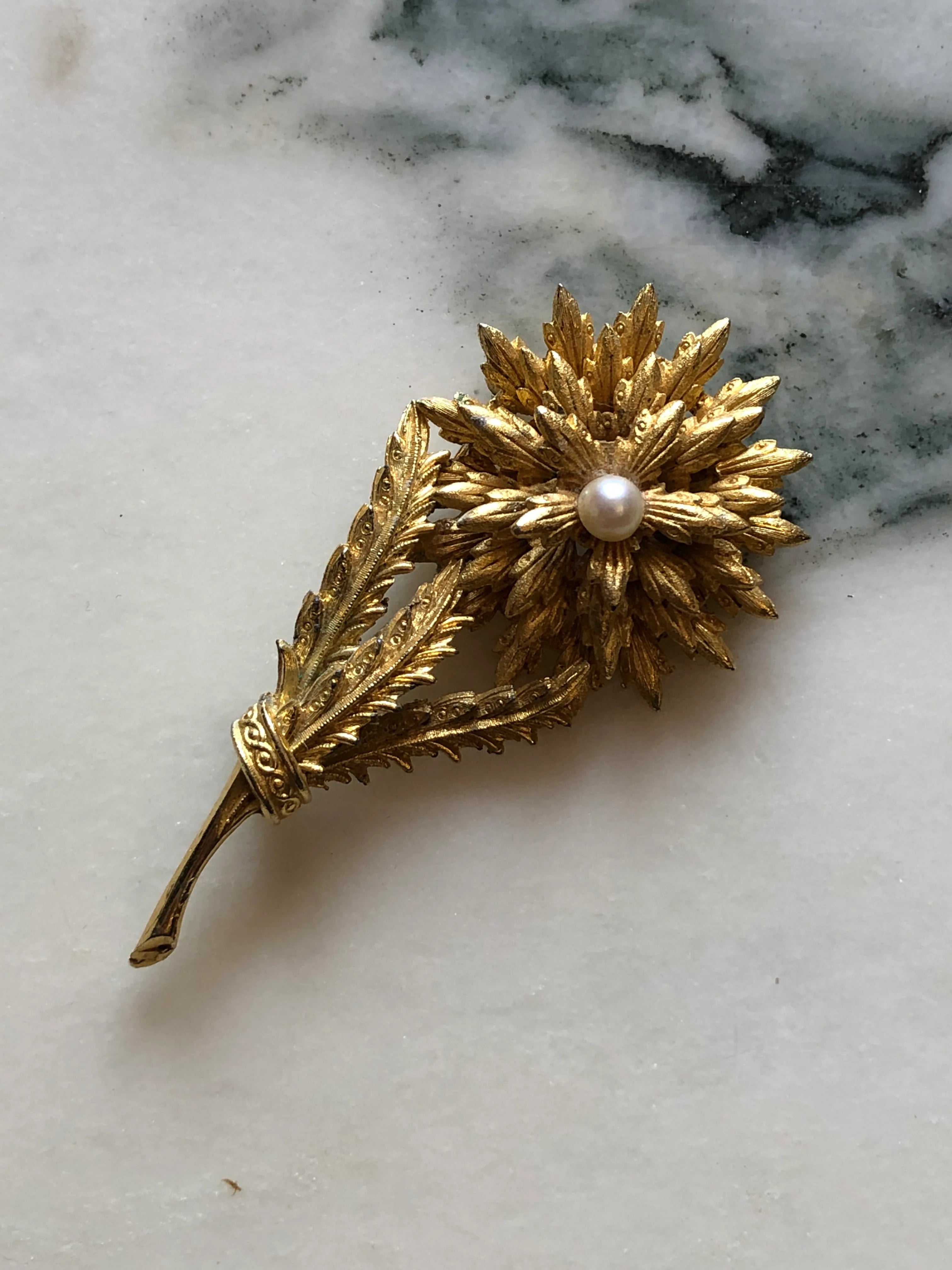 Broochella Perla d’Oro vintage gold-tone flower brooch with textured leaves, layered petals and a faux pearl centre. Pictured on a white marble surface. 