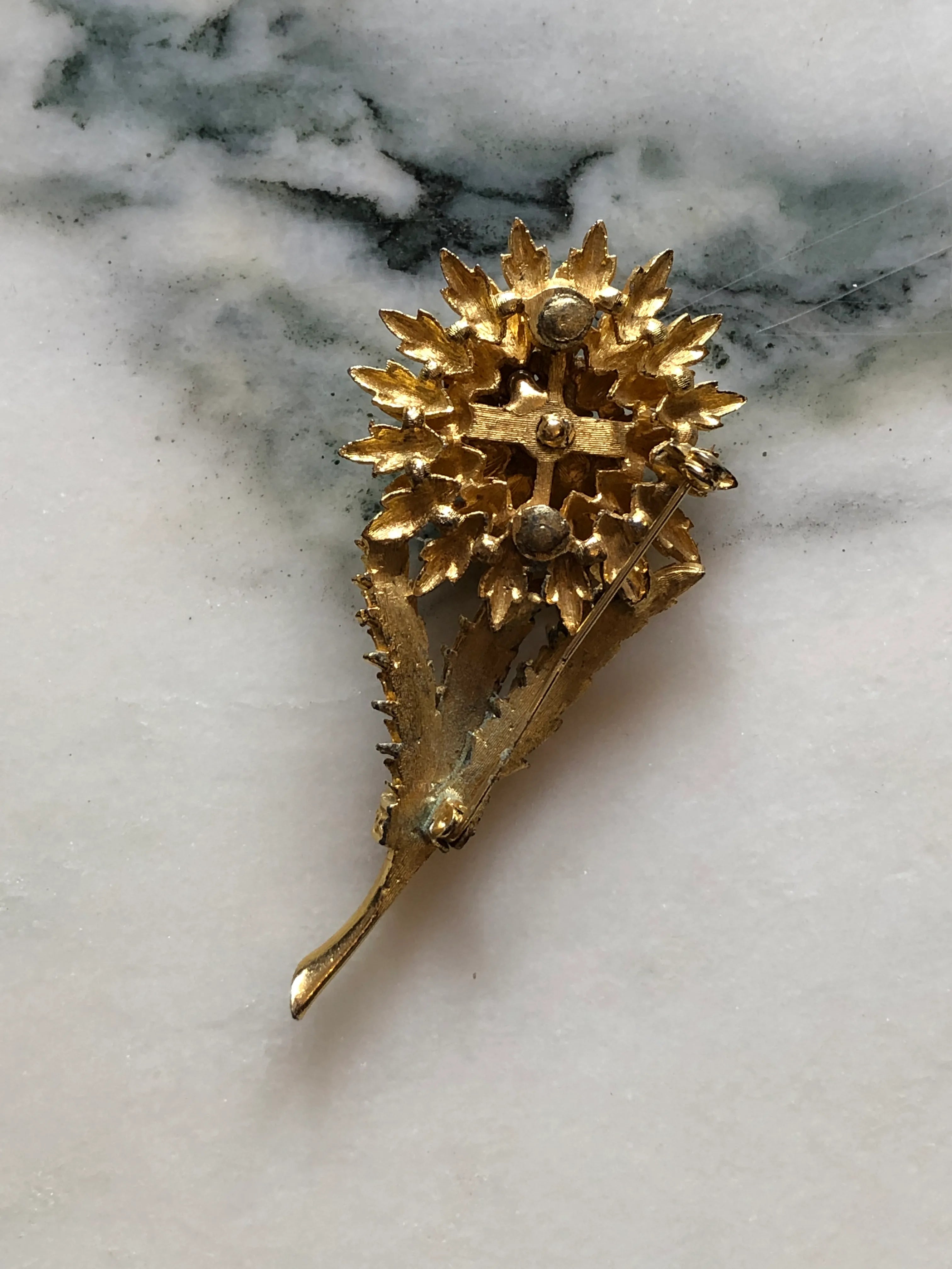 The back view of Broochella Perla d’Oro vintage gold-tone flower brooch with textured leaves, layered petals and a faux pearl centre. Pictured on a white marble surface. 