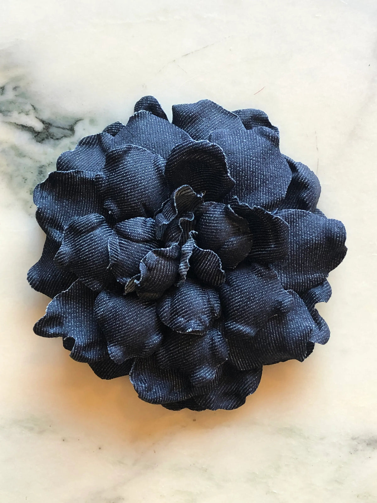 Broochella Pervinca Denim Flower Brooch in navy blue denim, featuring full layered sculptural petals with softly curved edges on a cream marble background.
