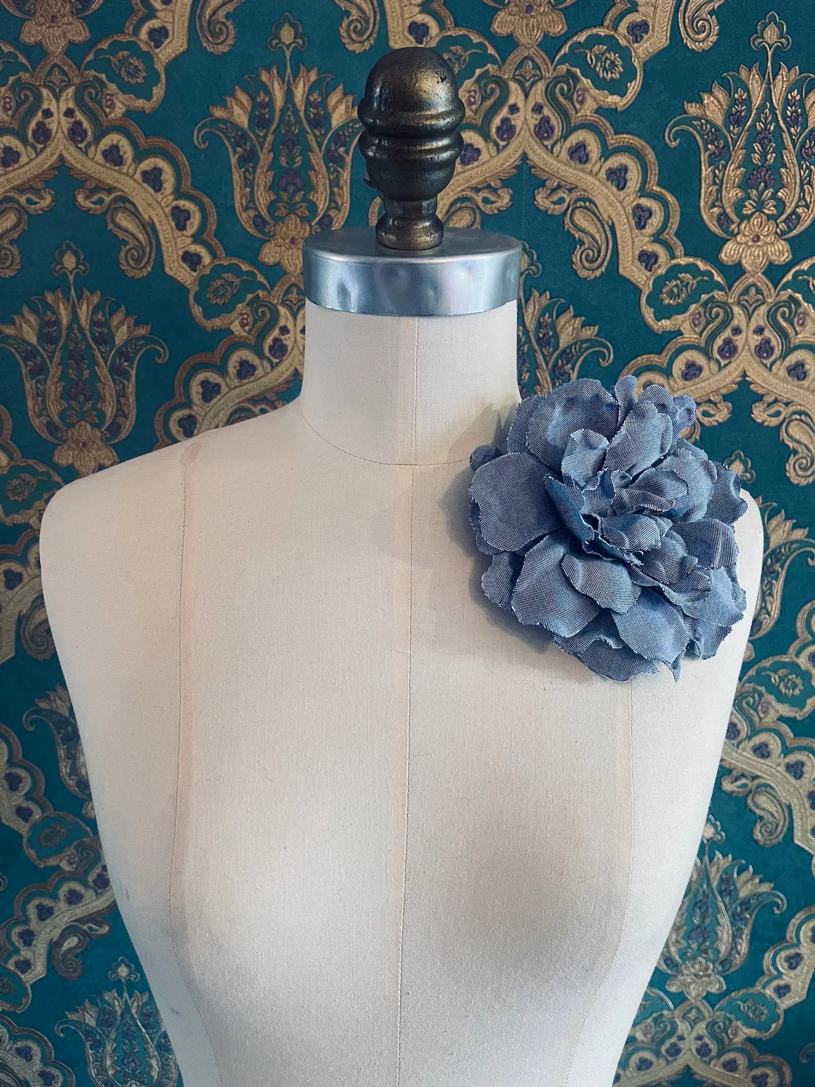 Broochella pervinca handmade blue denim flower brooch. Styled on a mannequin. The background is green and gold damask wallpaper