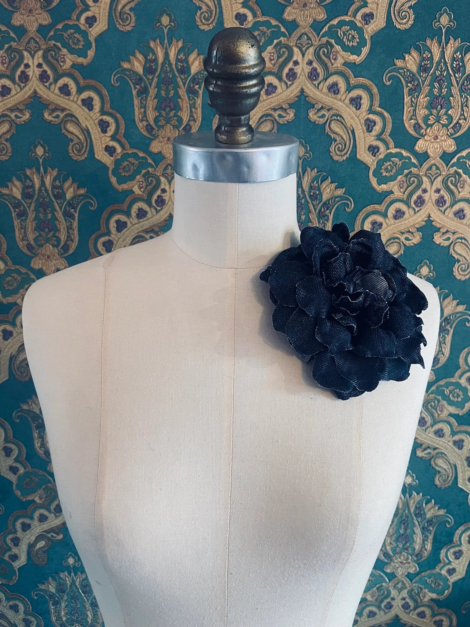 Broochella pervinca navy blue denim handmade flower brooch. Styled on a mannequin. The background is green and gold damask wallpaper
