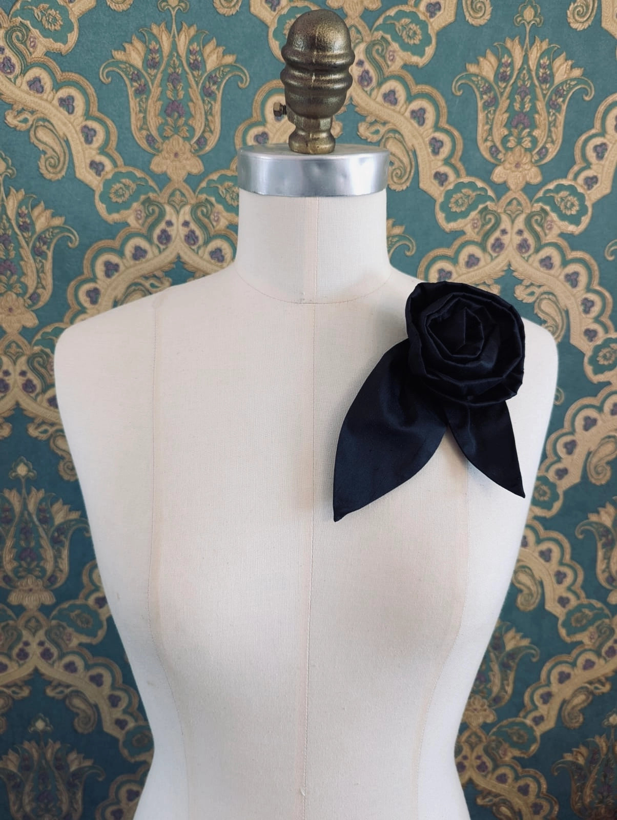 Broochella Petalo handmade silk flower brooch with petal detail in black. Pinned to a mannequin. The background is green and gold damask wallpaper.