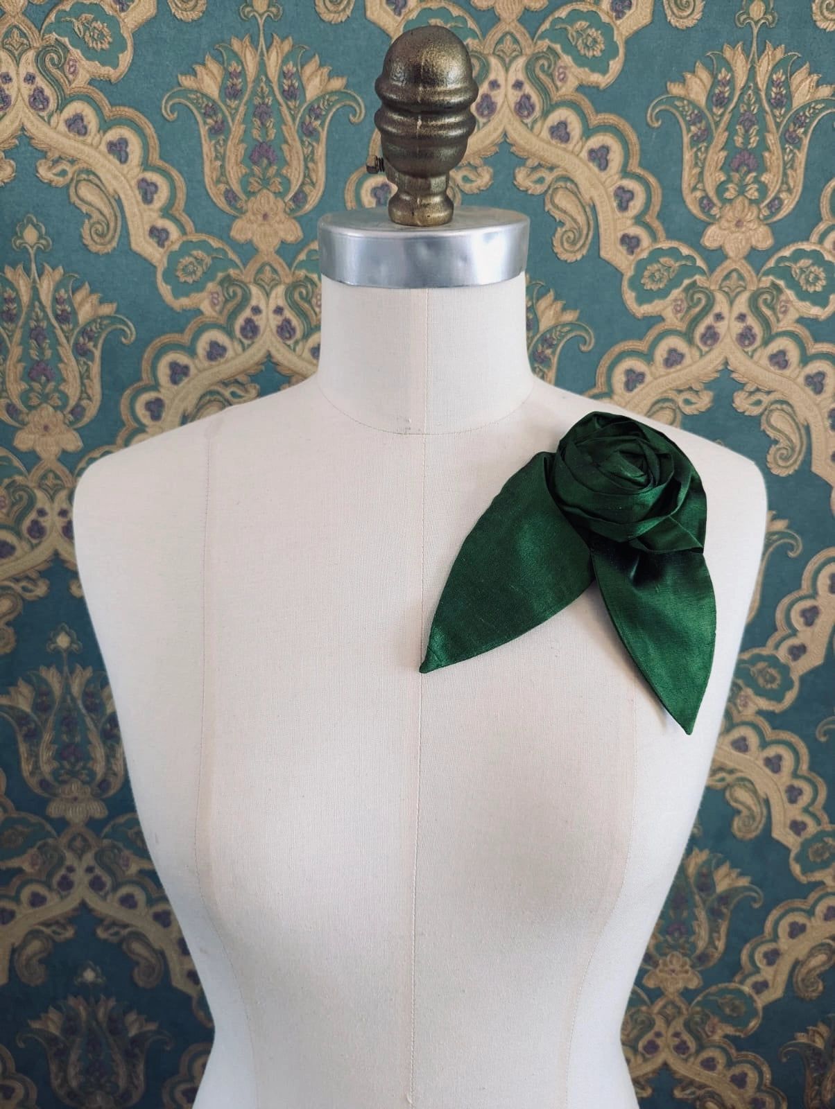 Broochella Petalo handmade silk flower brooch with petal detail in green. Pinned to a mannequin. The background is green and gold damask wallpaper.