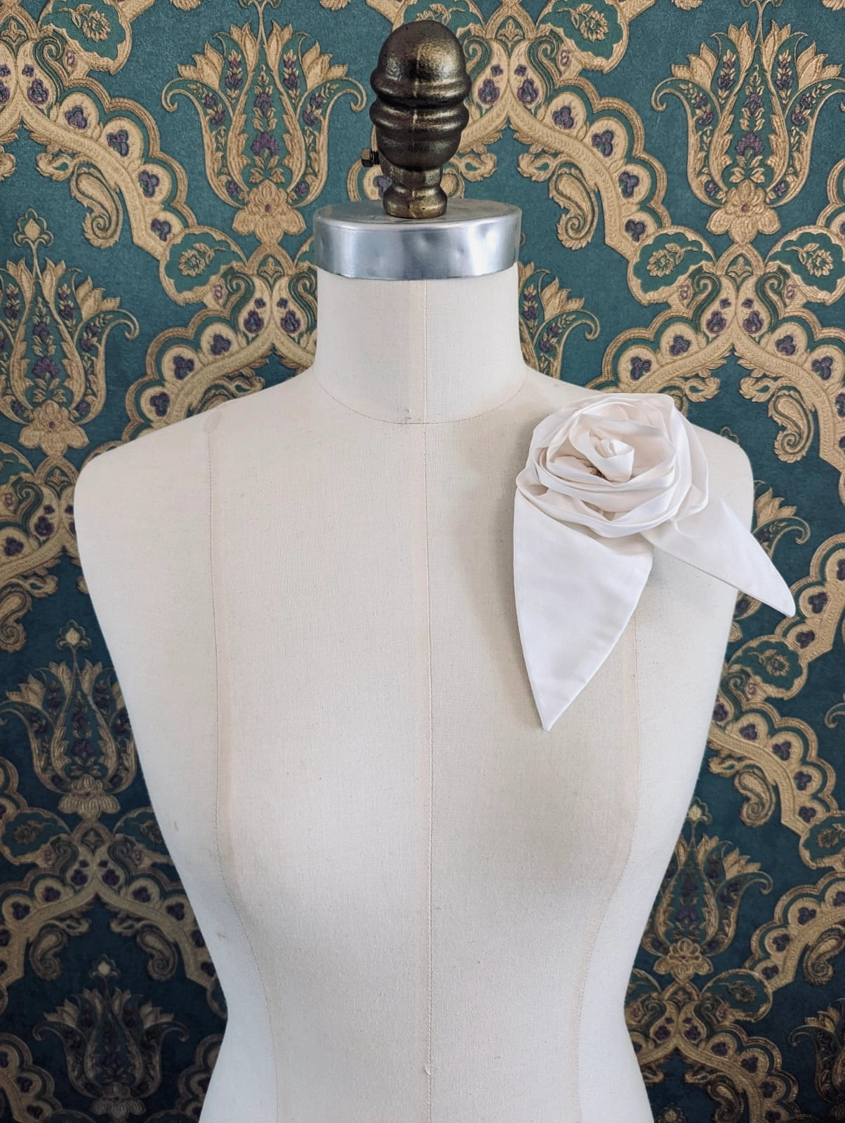 Broochella Petalo handmade silk flower brooch with petal detail in ivory. Pinned to a mannequin. The background is green and gold damask wallpaper.